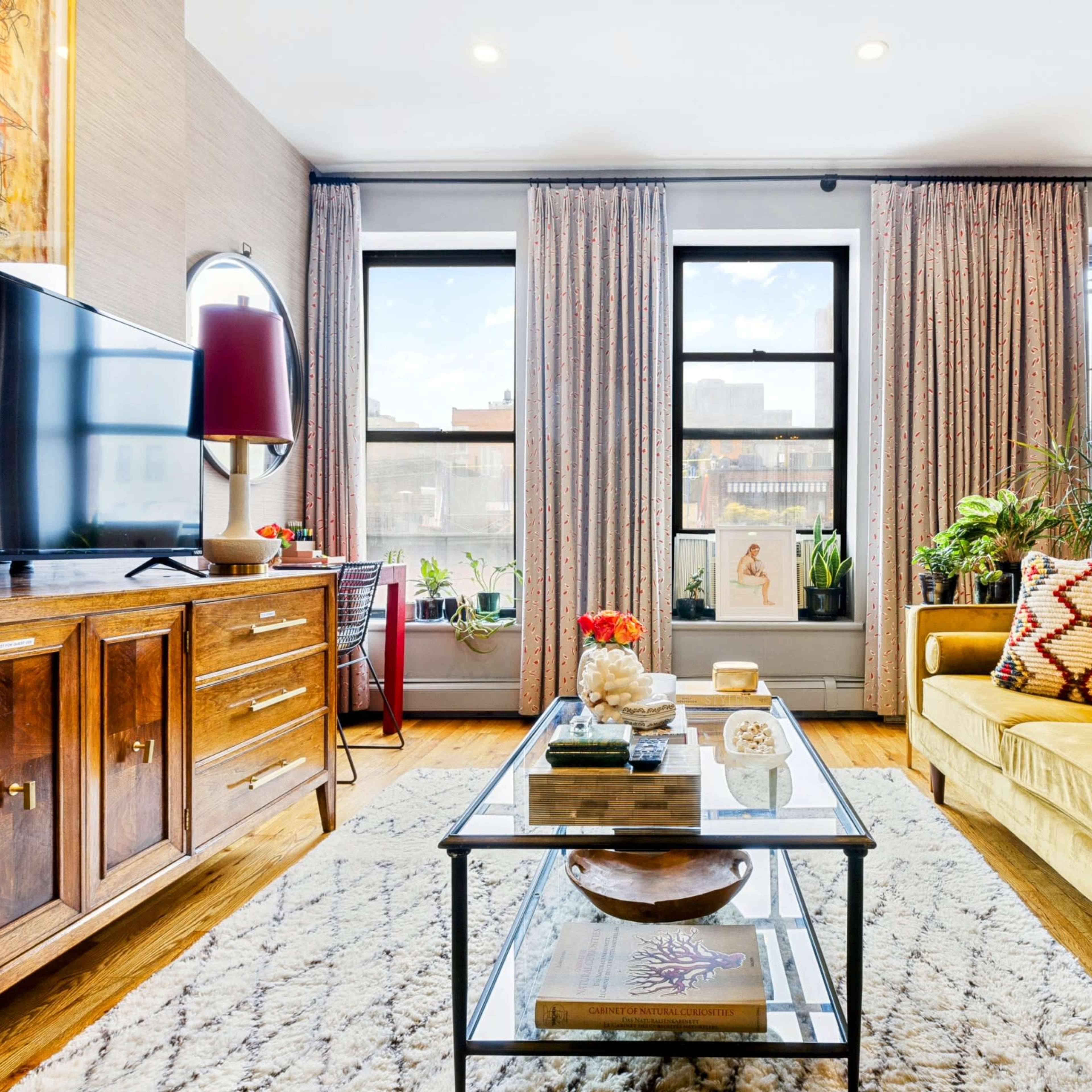 A living room features a yellow couch, a wooden television cabinet, sheer curtains, and a coffee table surrounded by plants and decorative items.
