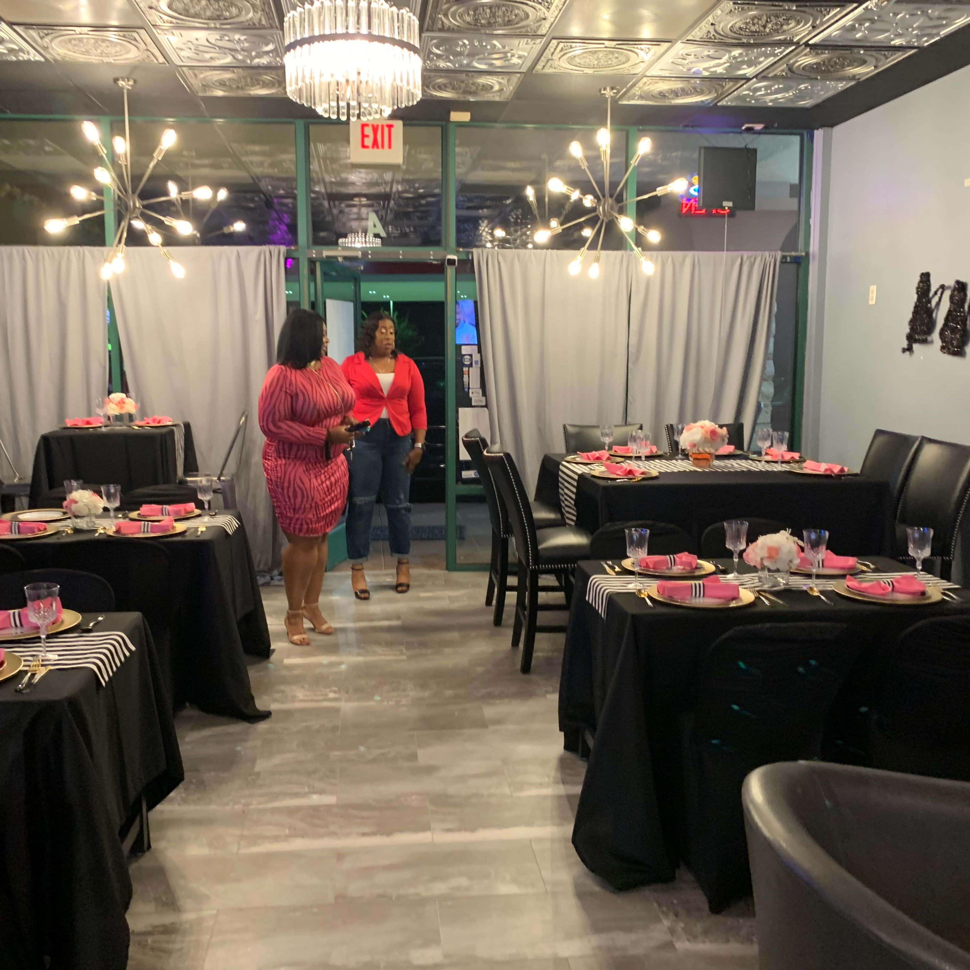 A modern dining space features several tables set with black tablecloths and pink dishware, while two women converse near the entrance.