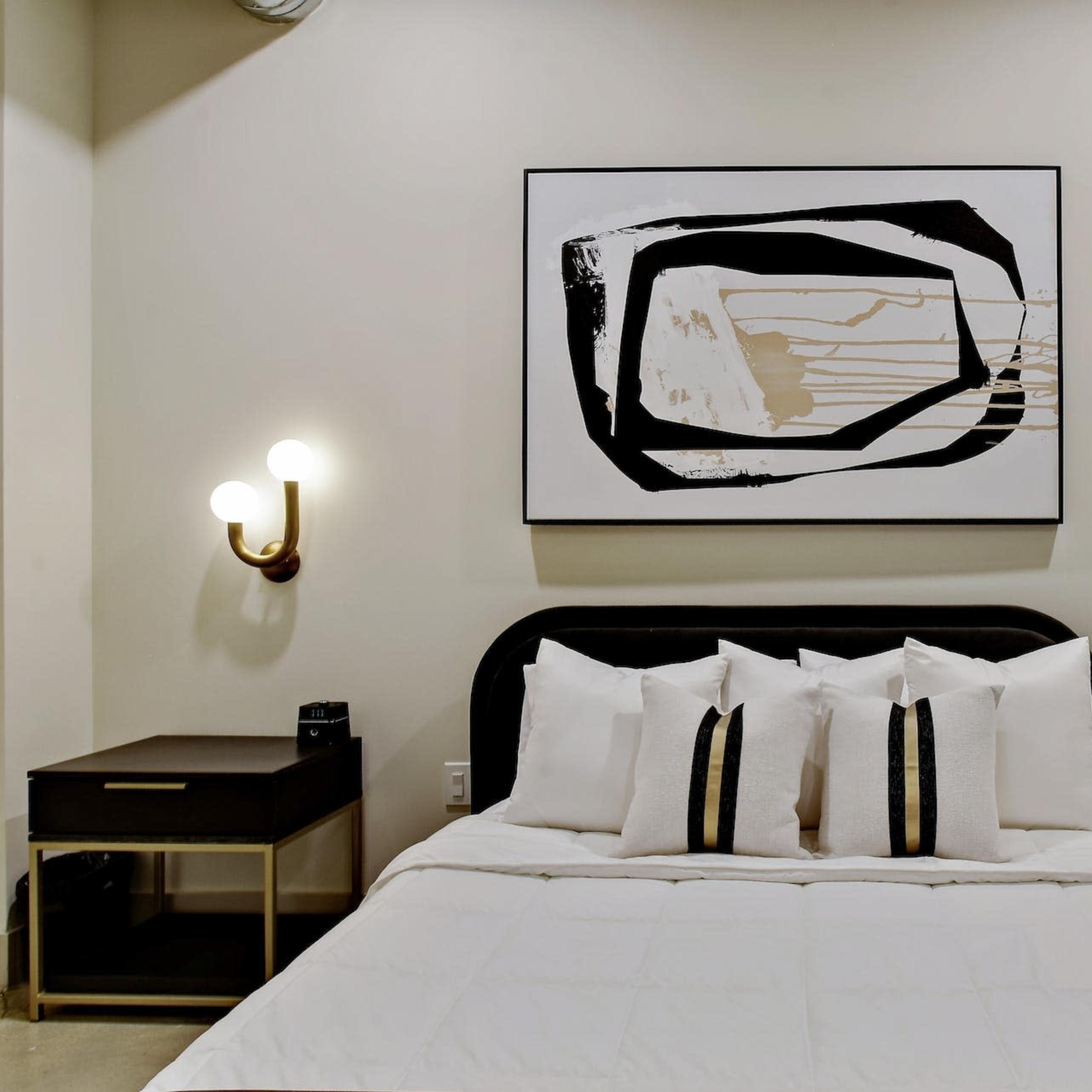 A bedroom with a bed featuring white pillows and a black-and-white abstract painting on the wall above it, flanked by two wall-mounted lights and bedside tables.