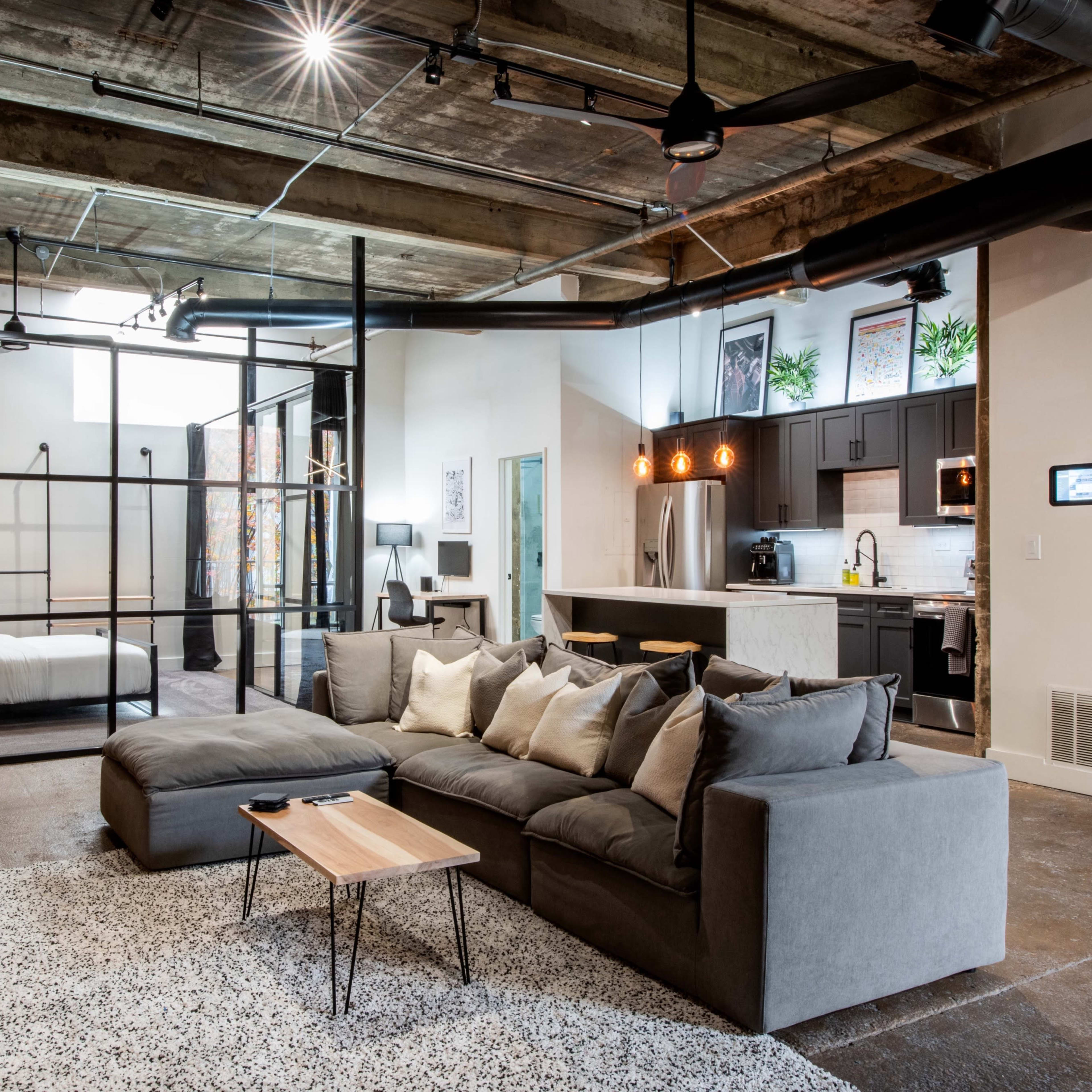 An industrial-style open-concept living space features a gray sectional sofa, a wooden coffee table, and visible kitchen and bedroom areas separated by glass partitions.