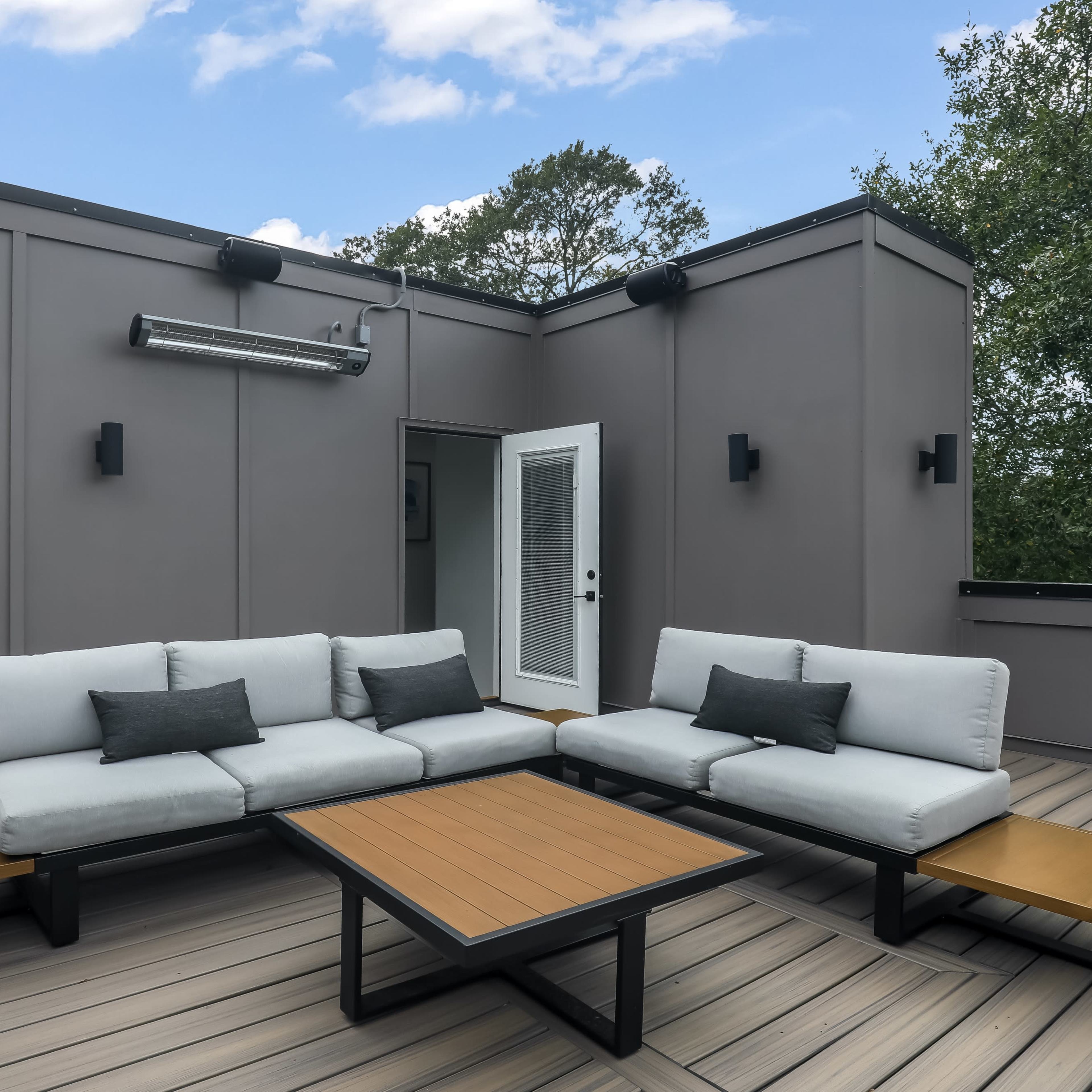 A modern rooftop terrace features a light gray sectional sofa, a wooden coffee table, and a doorway leading back inside.