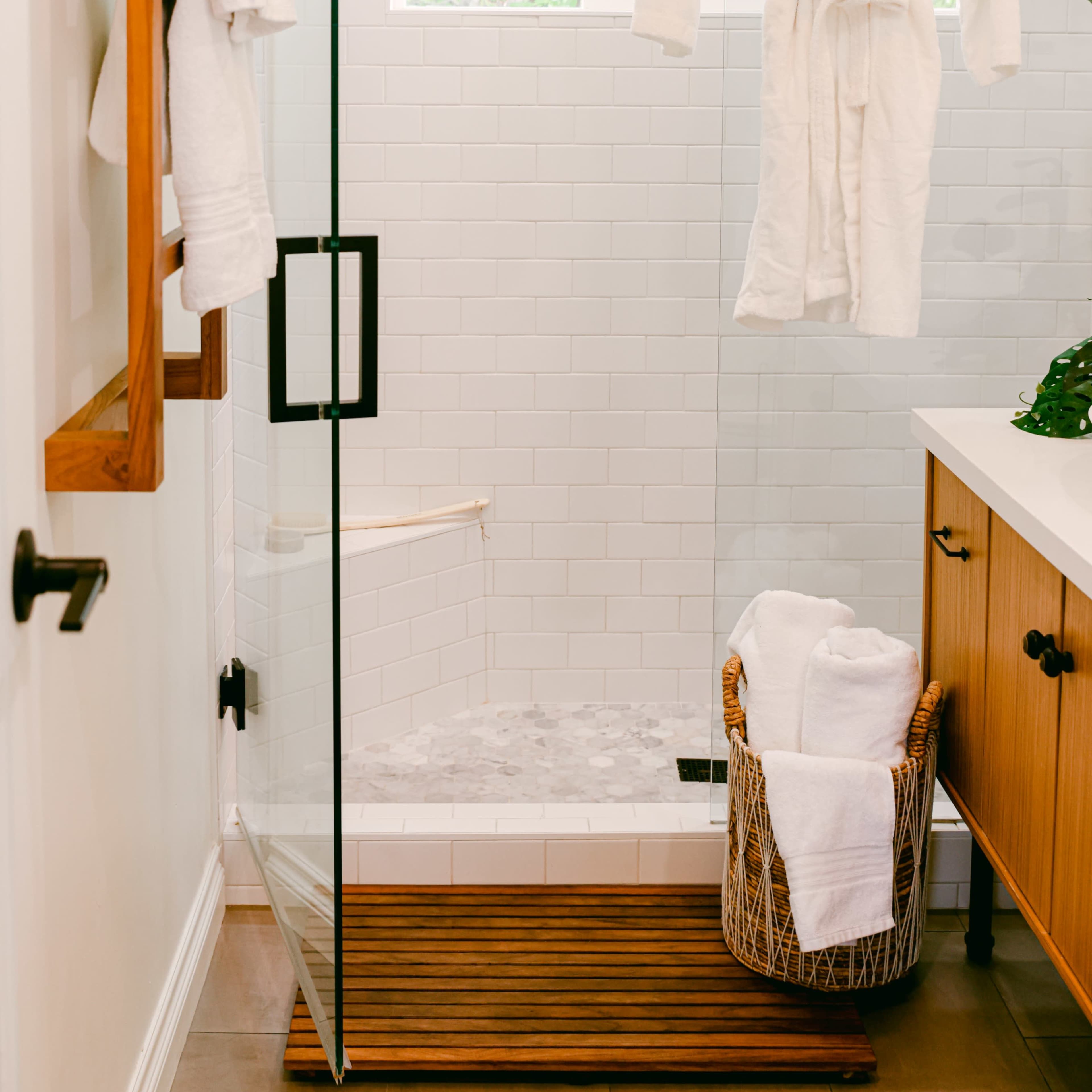 A glass shower enclosed by a wooden mat features a hanging white robe, with folded towels in a basket nearby and greenery visible outside a window.
