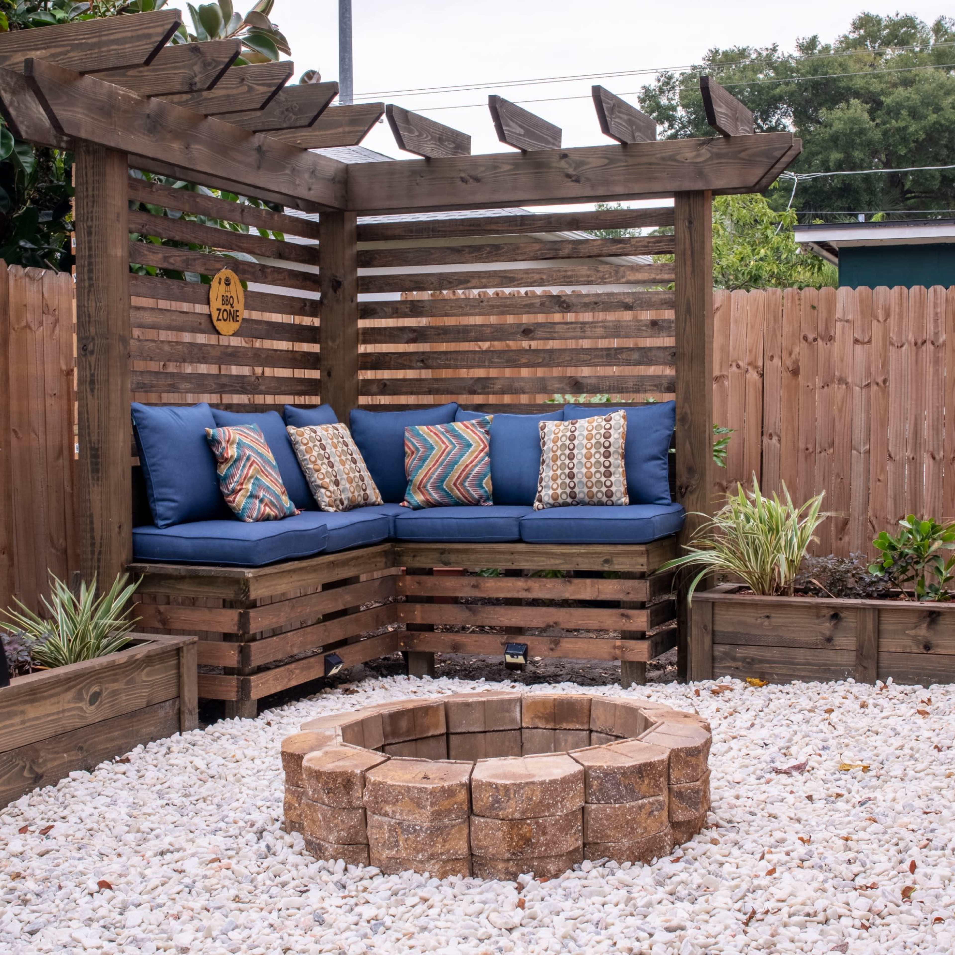 A wooden outdoor seating area with a fire pit surrounded by white gravel and bordered by a wooden fence.