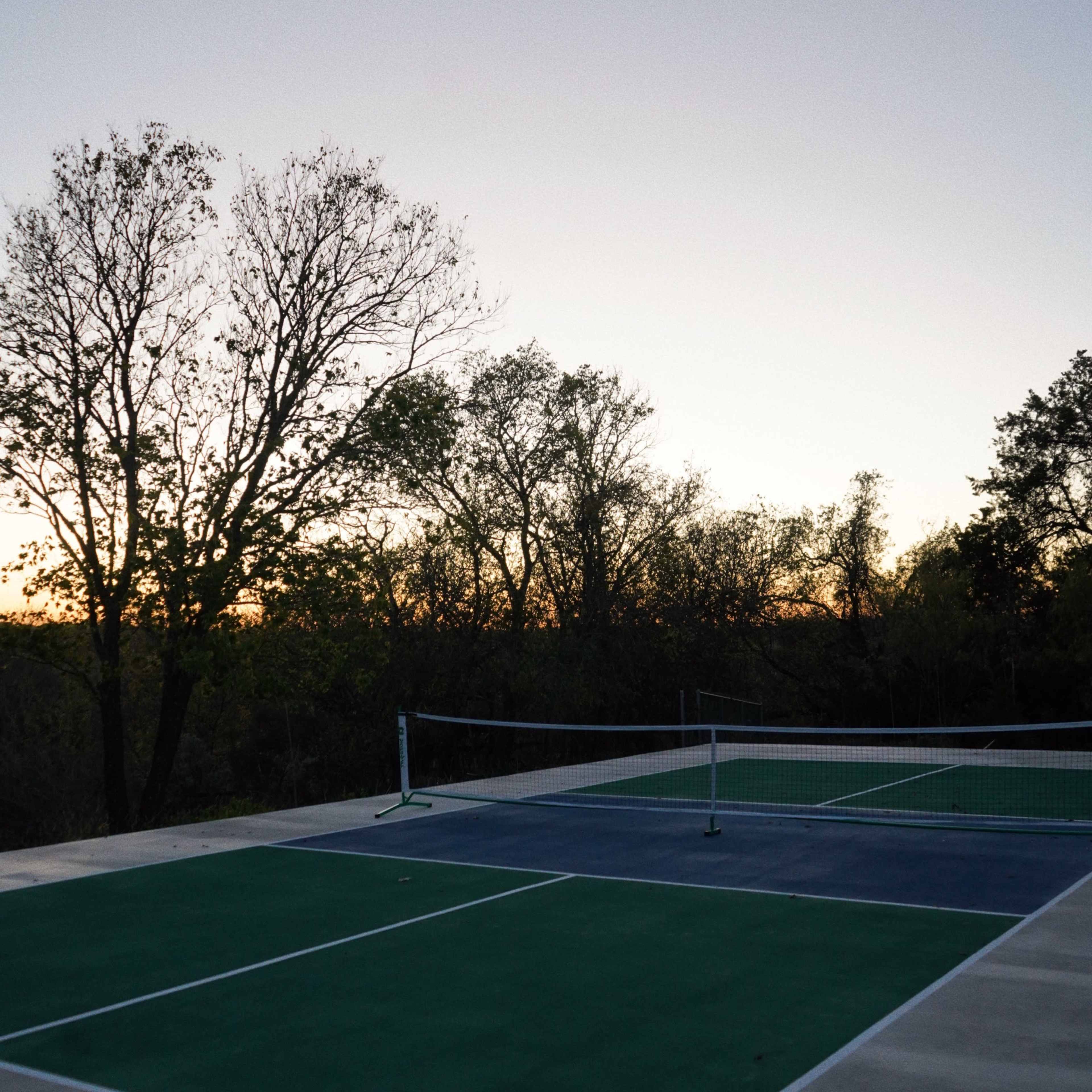 A tennis court with green and blue surfaces is visible at dusk, surrounded by trees.