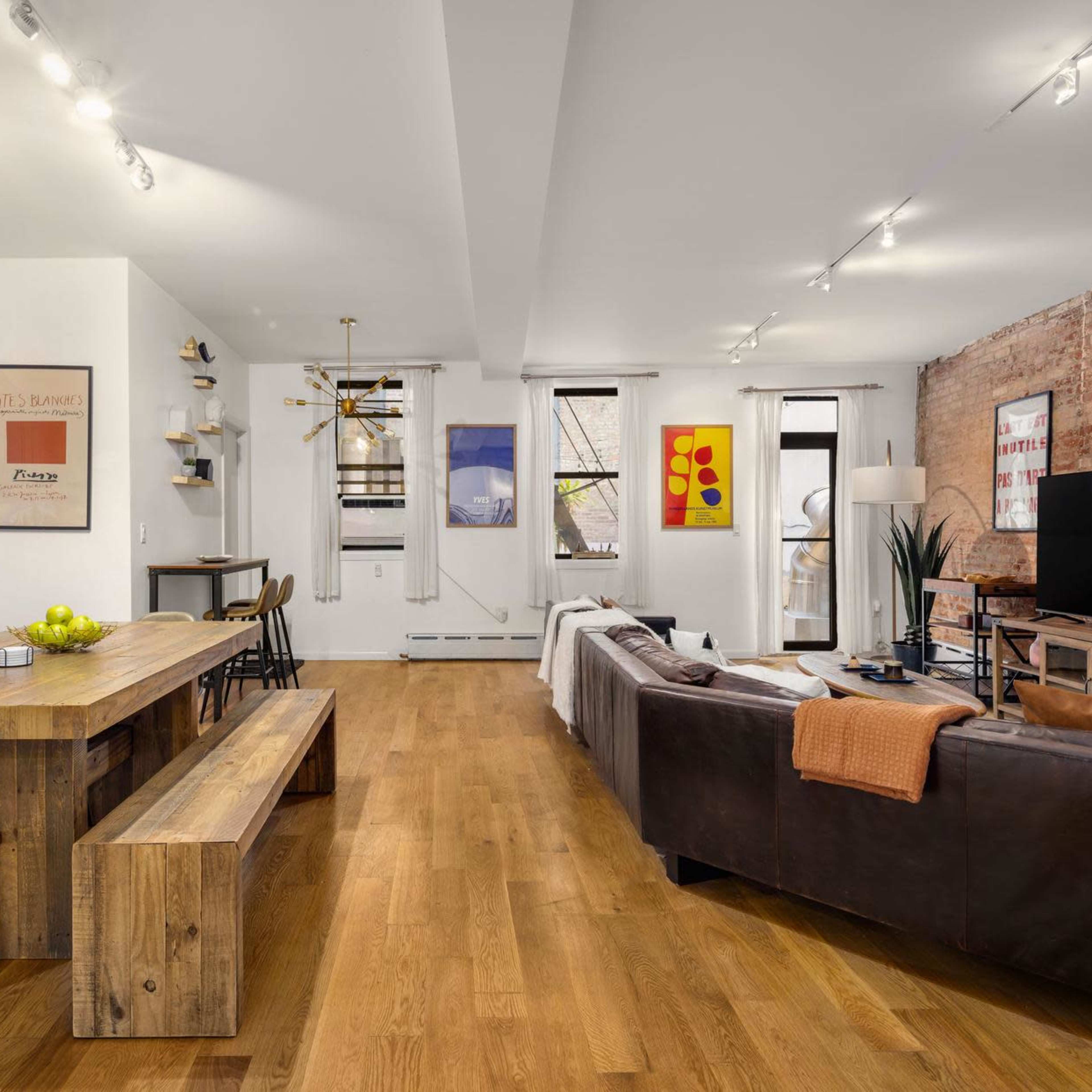 A modern living space featuring a kitchen area with gray cabinets, a wooden dining table, a brown sofa, and exposed brick walls.