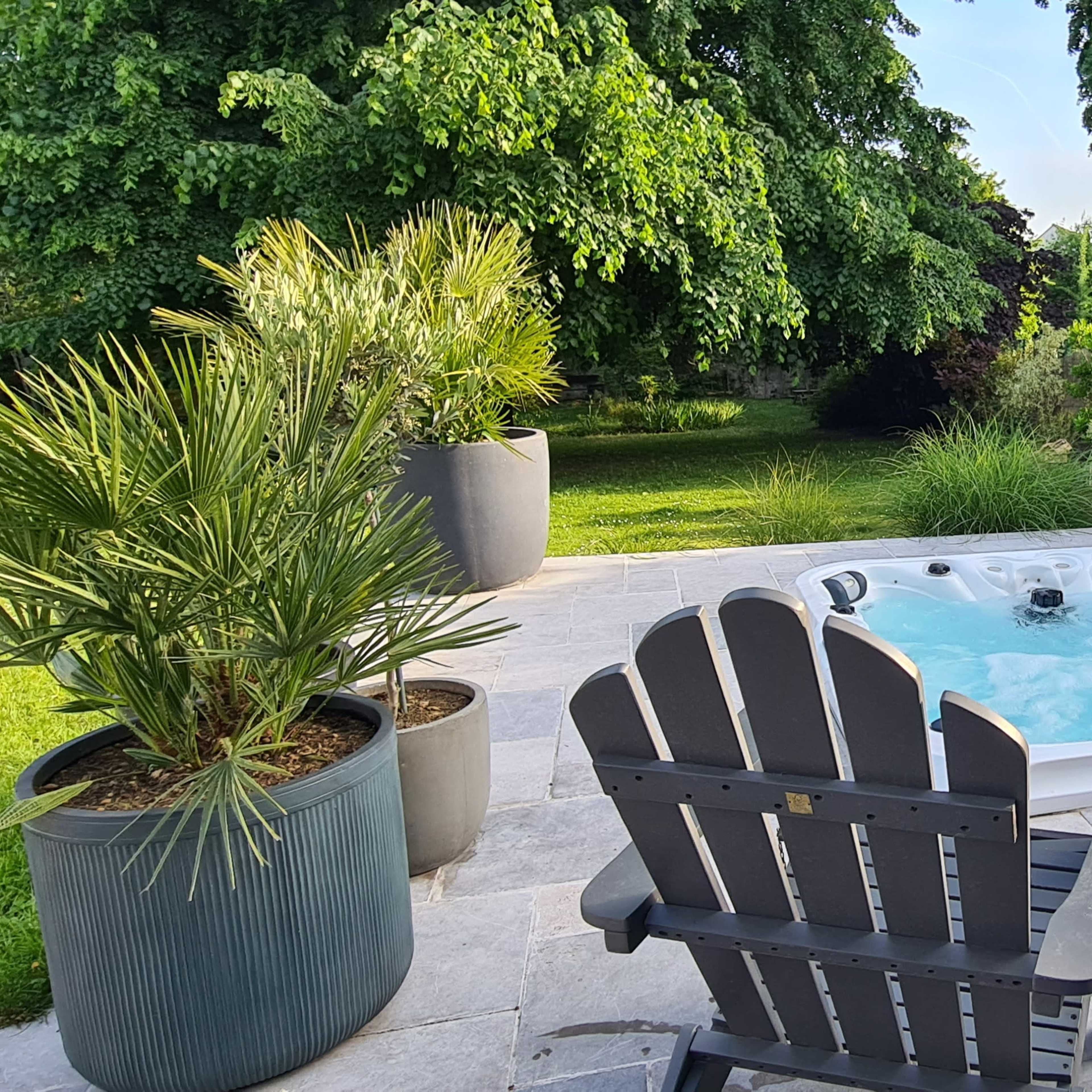 A hot tub is situated on a stone patio surrounded by green plants and trees.