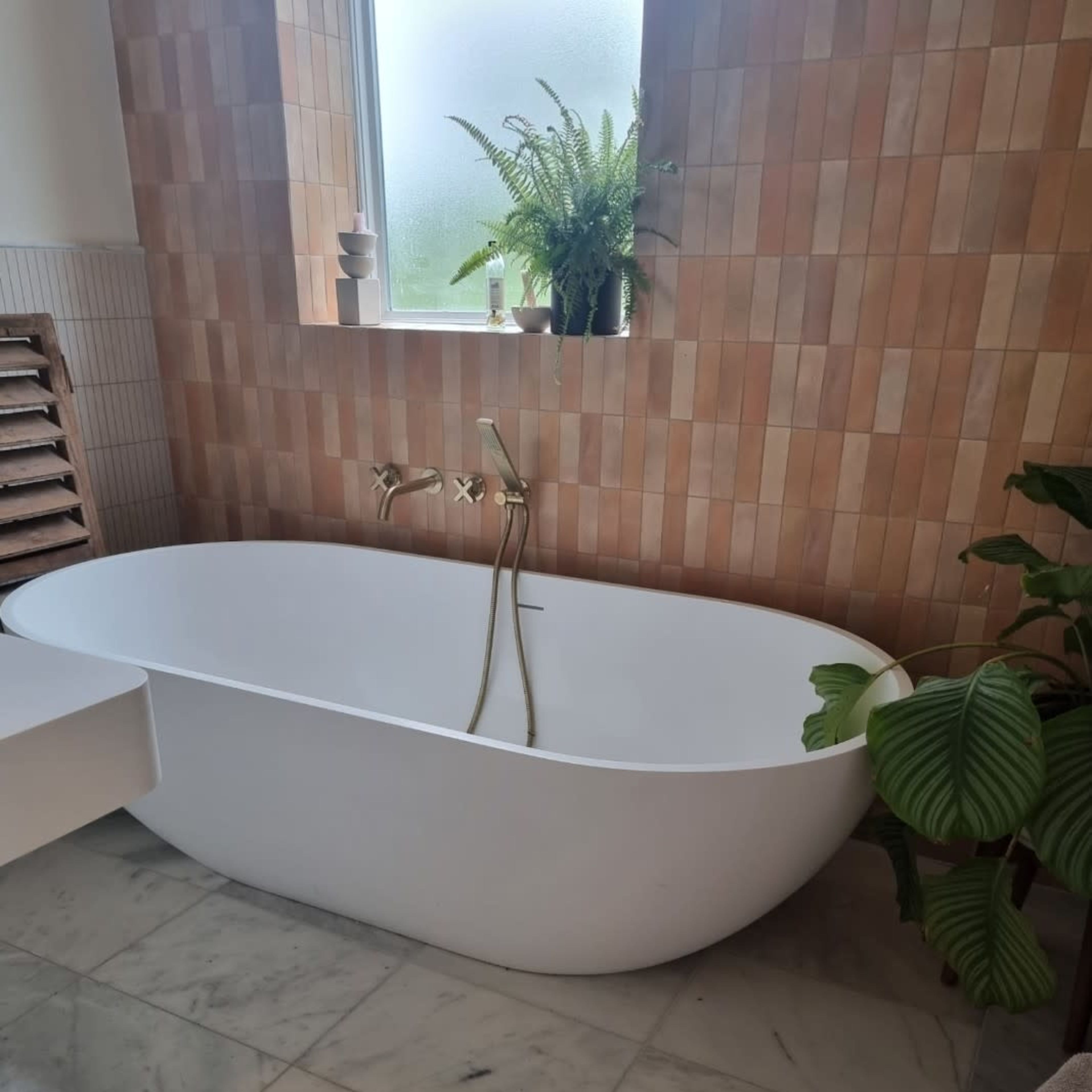 A freestanding white bathtub surrounded by tiled walls and greenery in a bathroom setting.