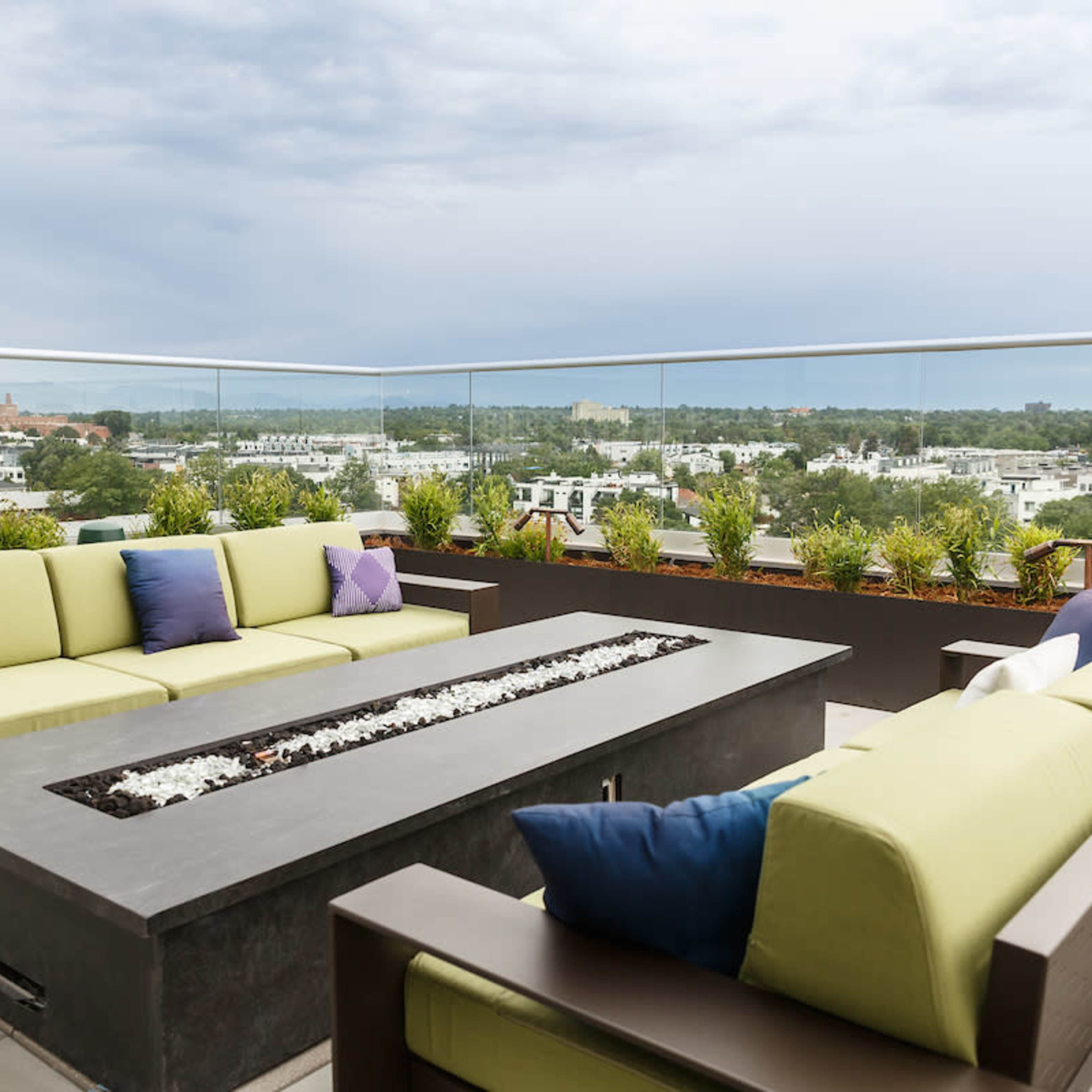A rooftop terrace features modern seating with green cushions and a rectangular fire pit, surrounded by planters and a view of the city below.