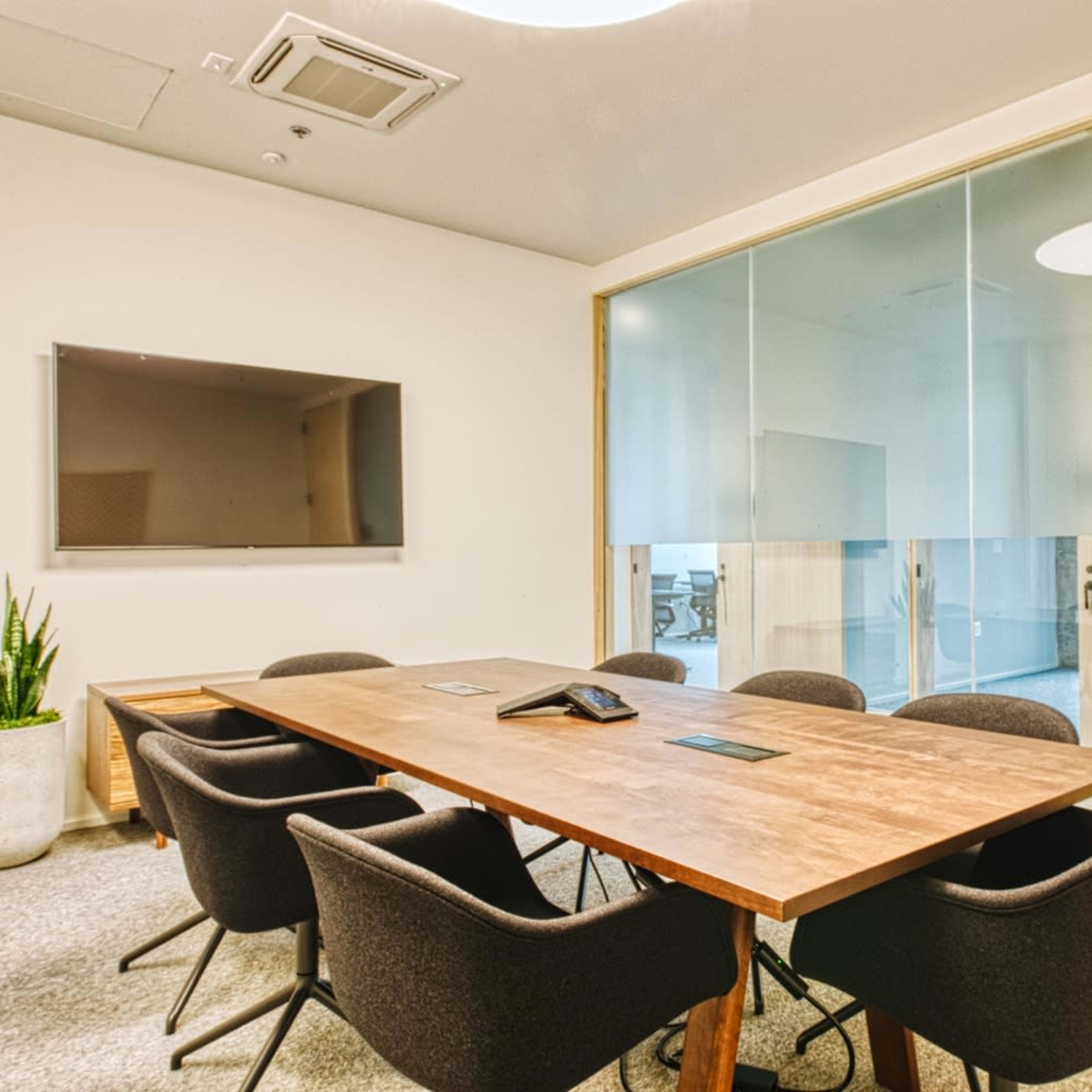 A modern conference room features a wooden table with seating for six, a large wall-mounted TV, and a glass wall separating it from another room.