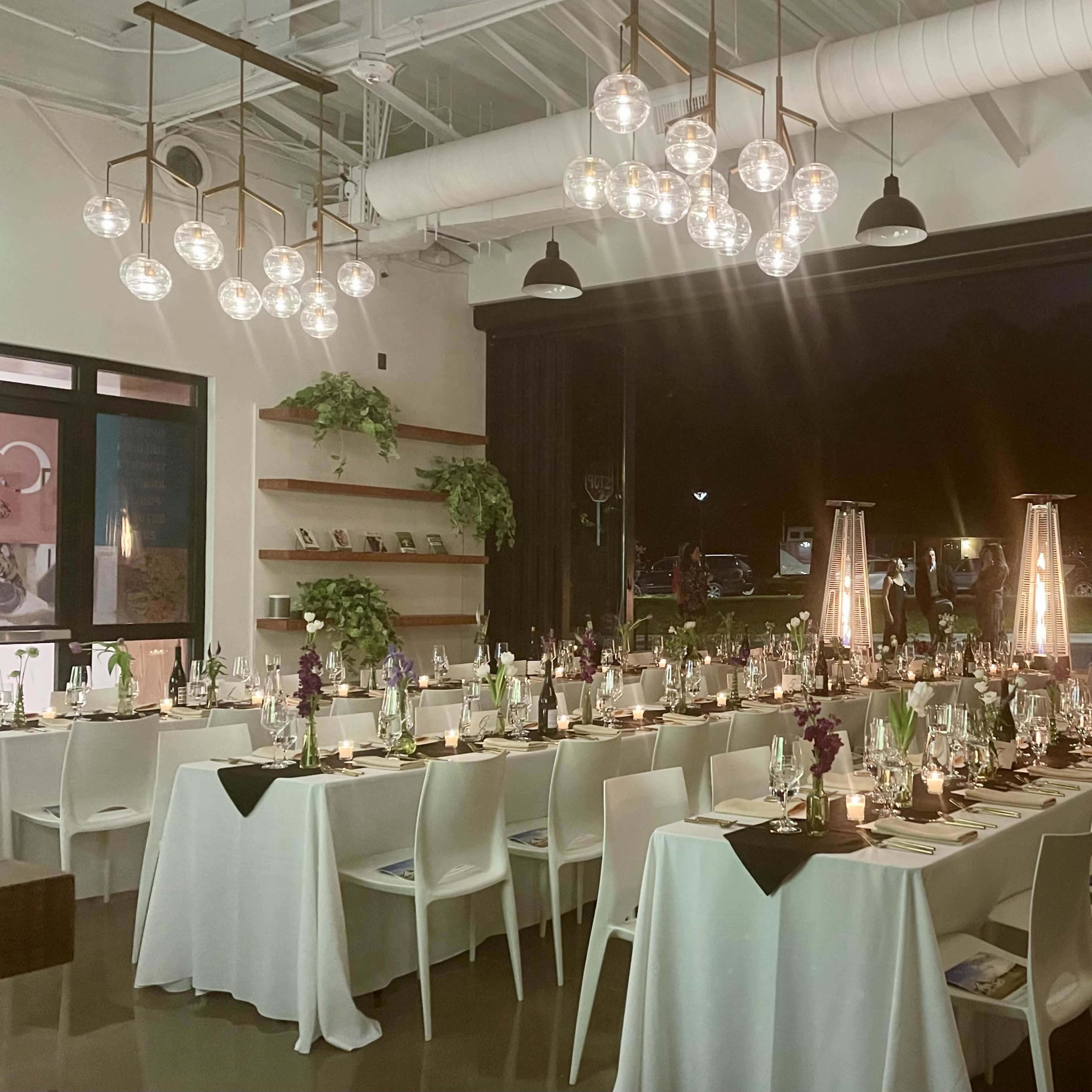 A long, elegantly set dinner table is arranged in a dimly lit modern venue, featuring glass pendant lights and candles on each table.
