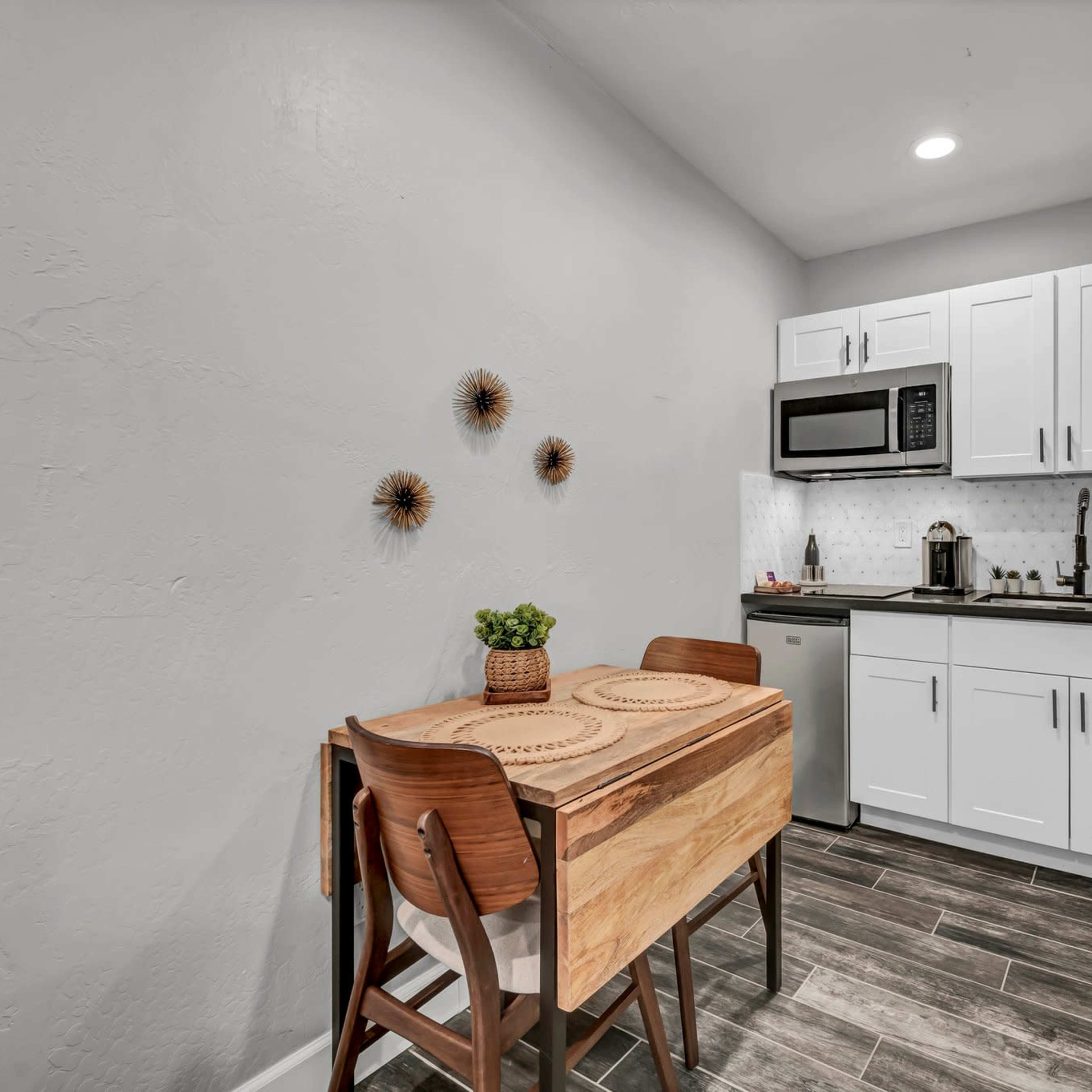 A compact kitchen features a small wooden dining table, stainless steel appliances, and light-colored cabinetry against a gray wall.
