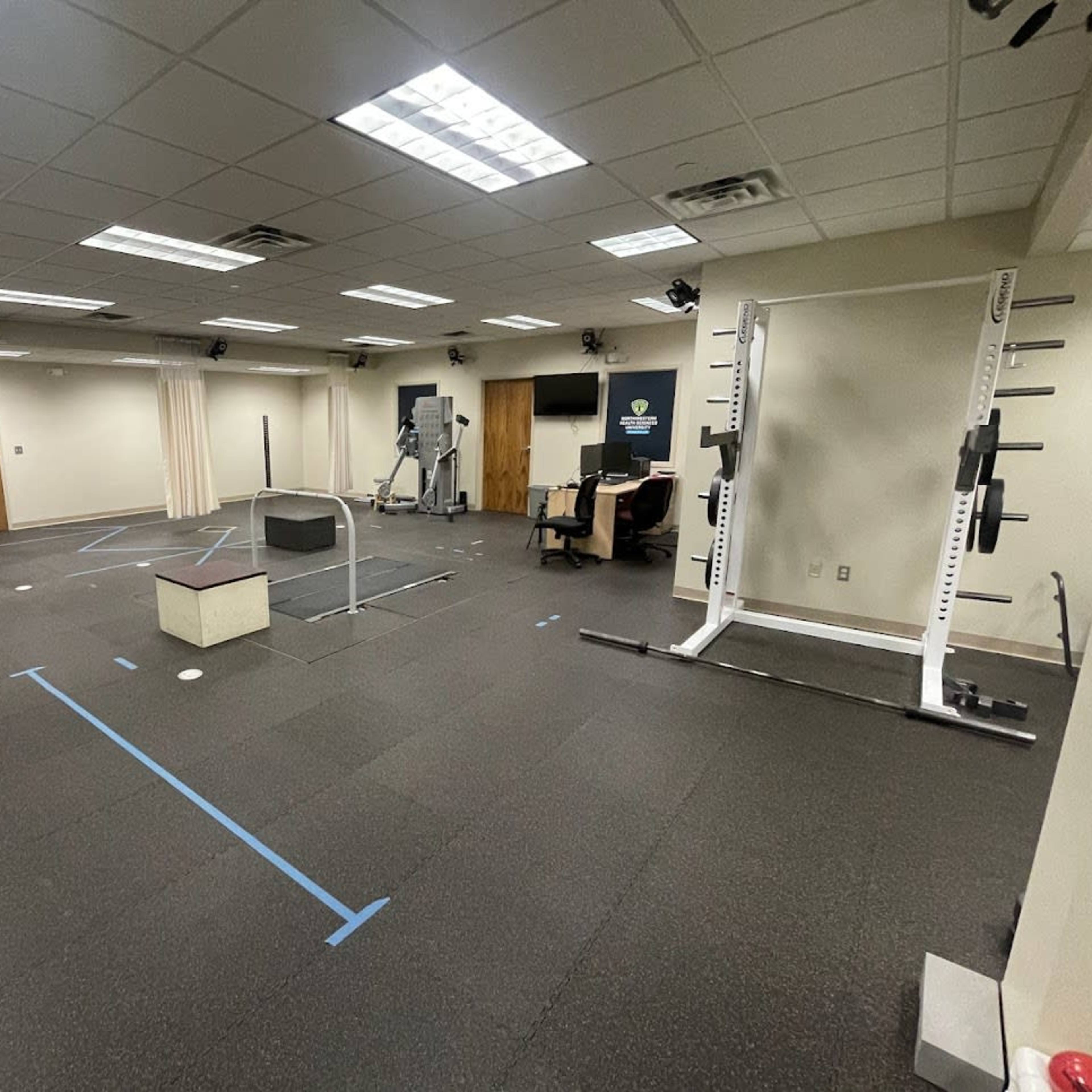 A gym interior with various exercise equipment, including a treadmill, a squat rack, and a plyometric box, arranged on a rubber floor.