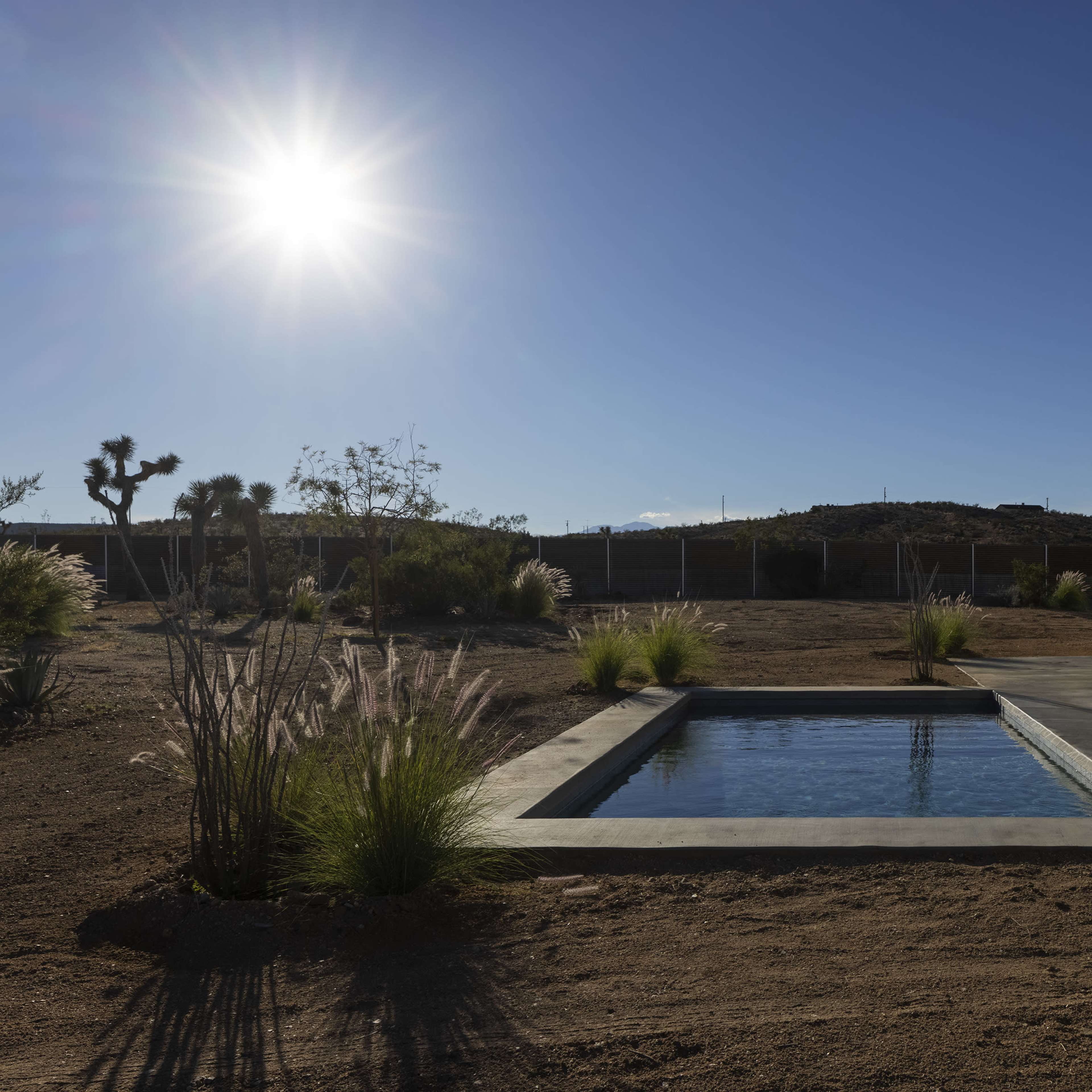 A modern house with a small pool is set in a desert landscape under a clear blue sky with the sun shining brightly.