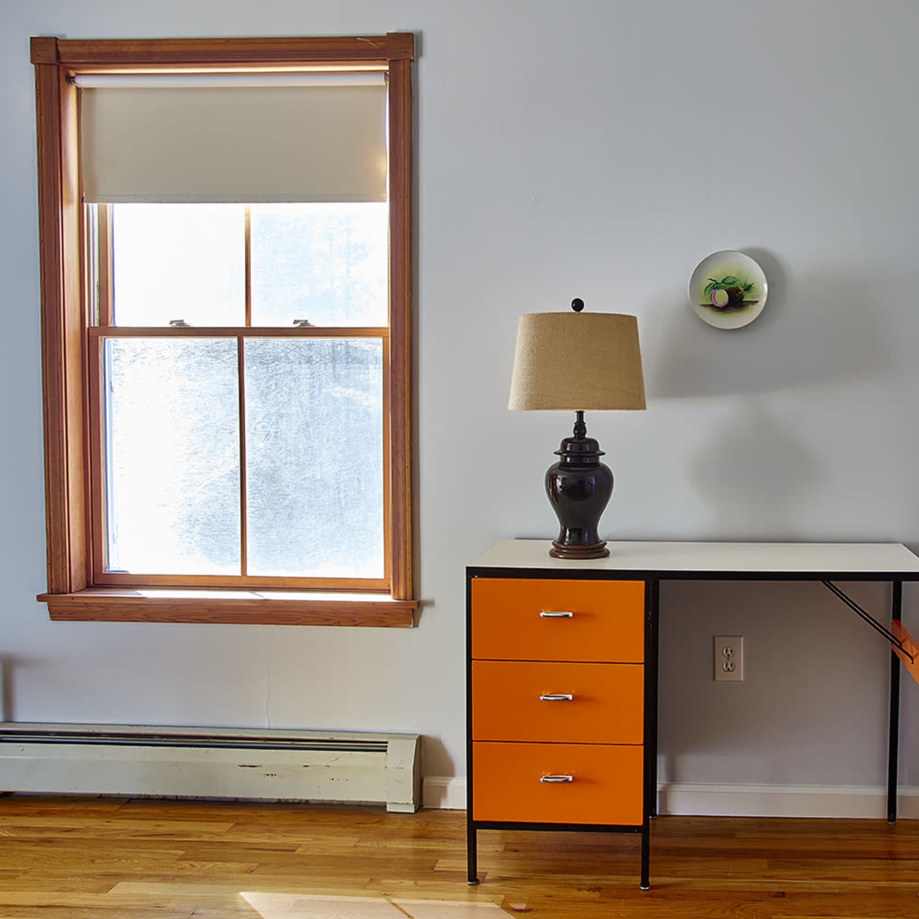A small desk with an orange drawer, a lamp, and a wall-mounted circular picture, situated beside a window with a blind.