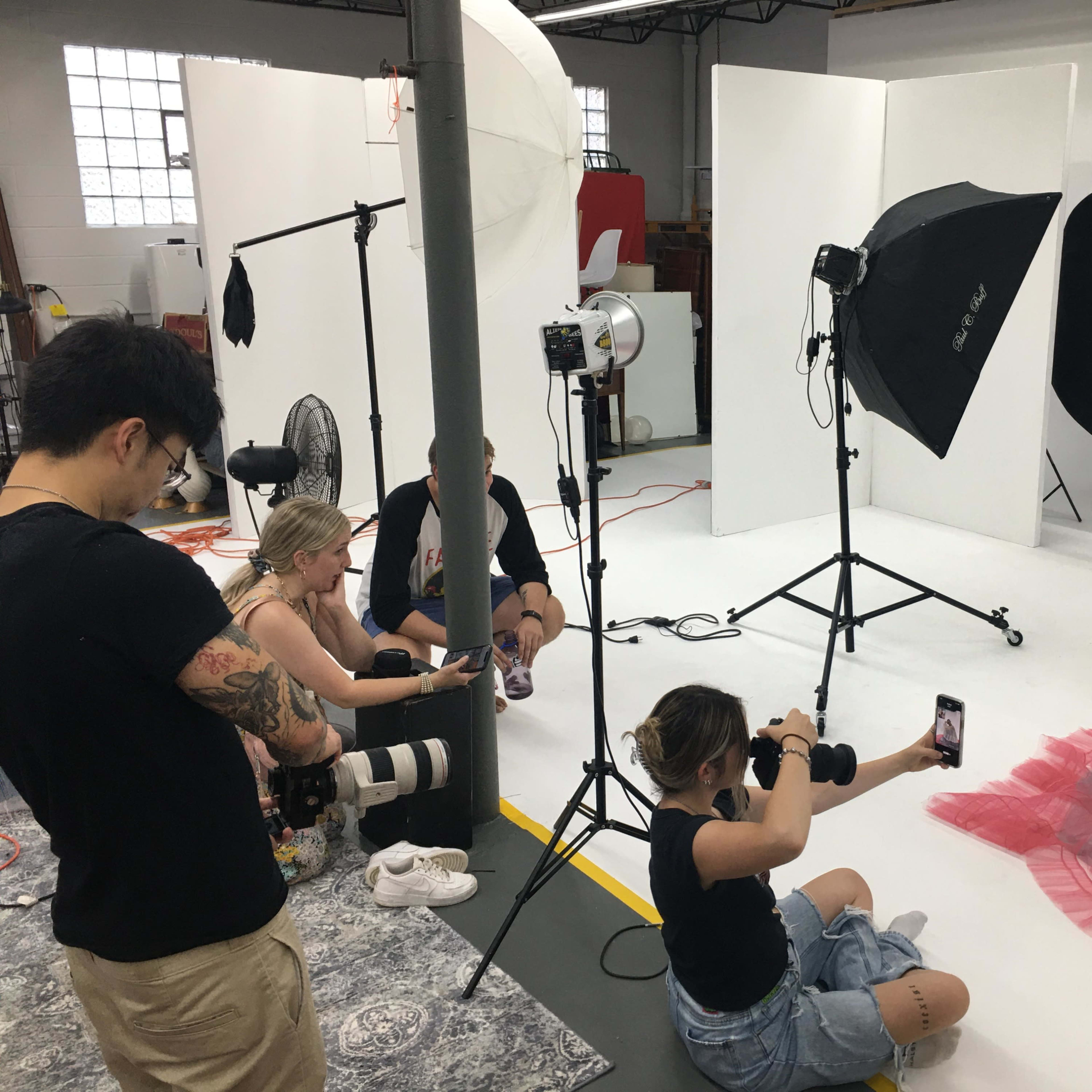 A group of photographers and assistants are setting up a photoshoot with a model in a studio, featuring backdrop lights and equipment.
