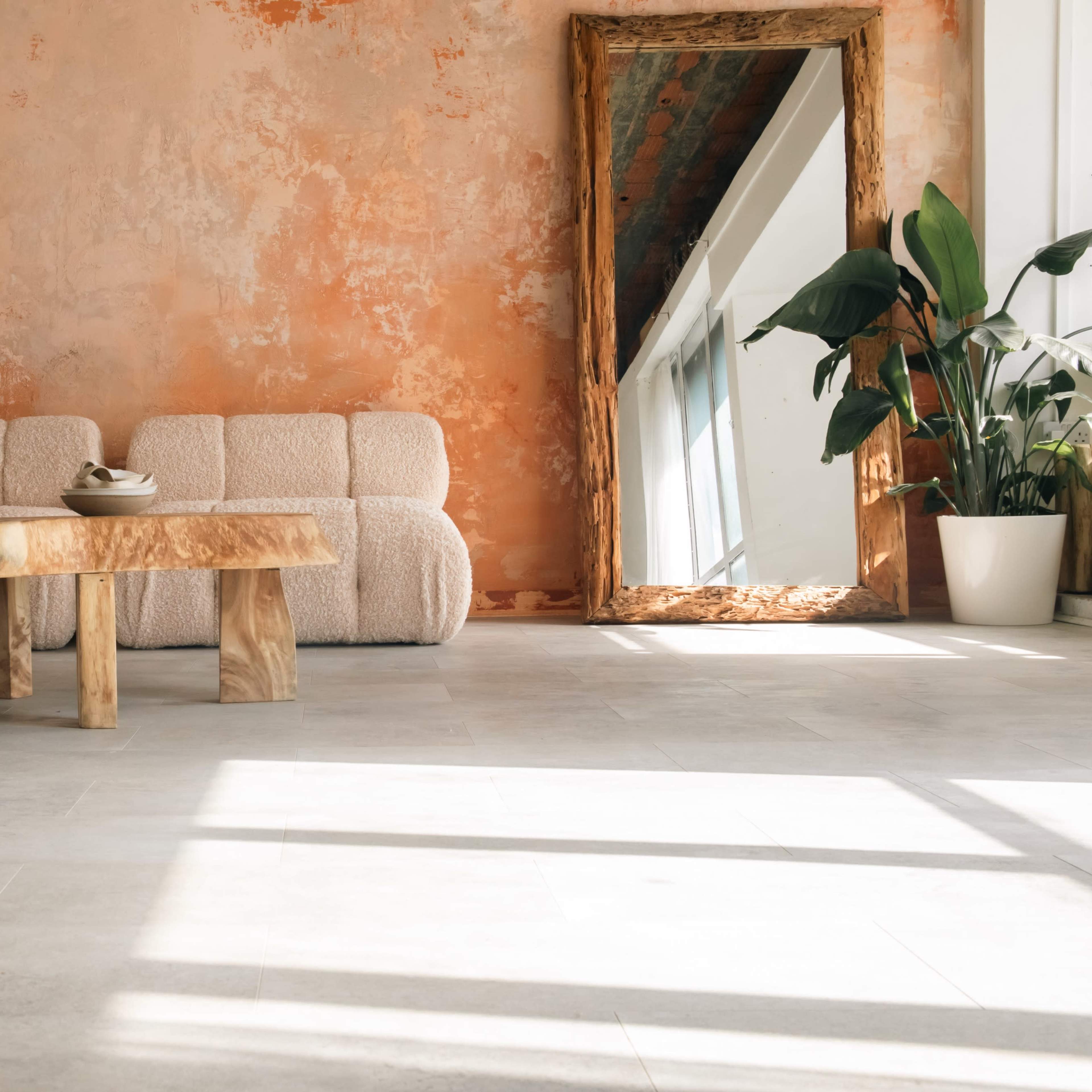 A cozy interior with a textured orange wall, a plush white sofa, a wooden table, and potted plants near a large mirror reflecting natural light.