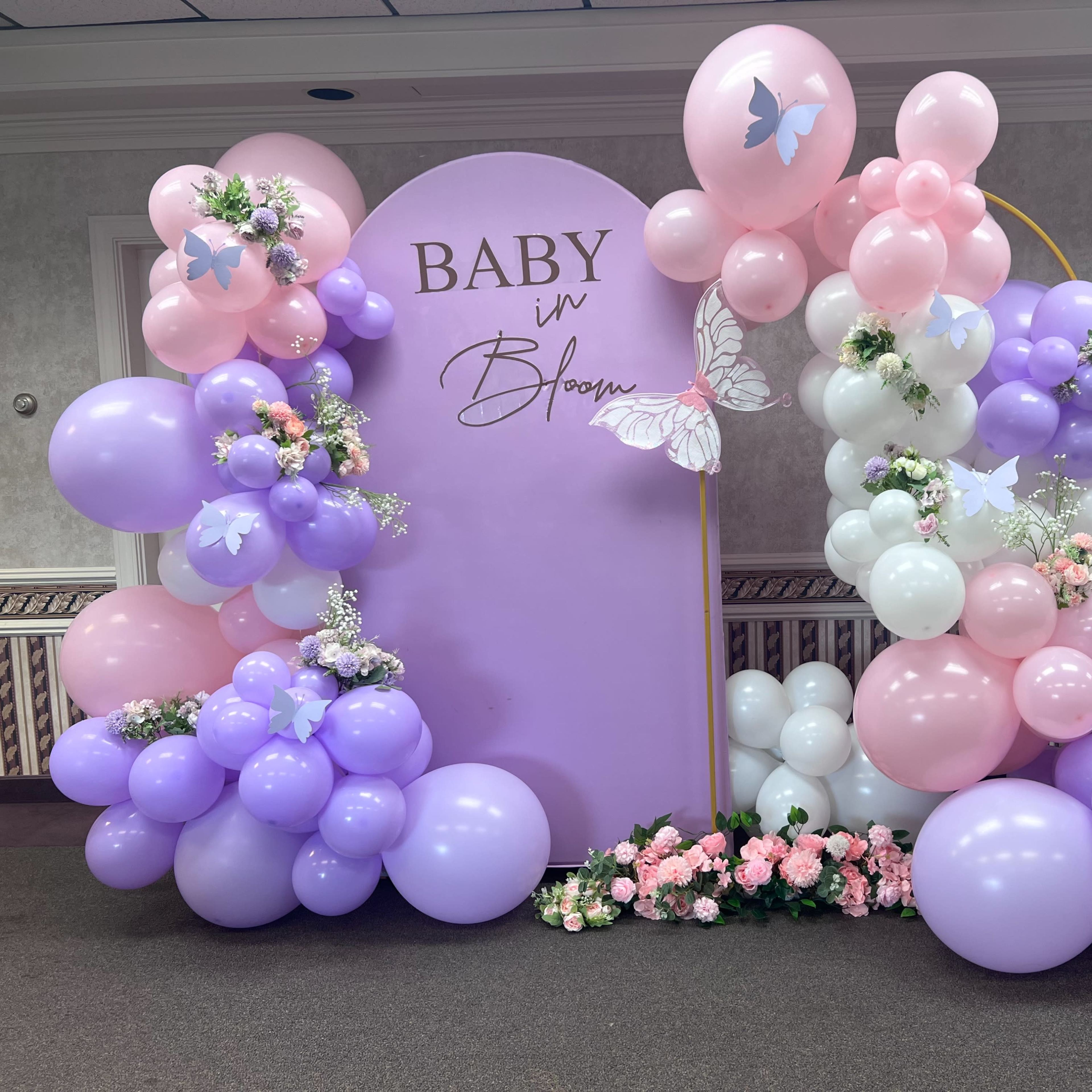 A decorative setup for a baby shower, with an arch made of purple, pink, and white balloons, surrounded by flowers and a central sign that reads "Baby in Bloom."