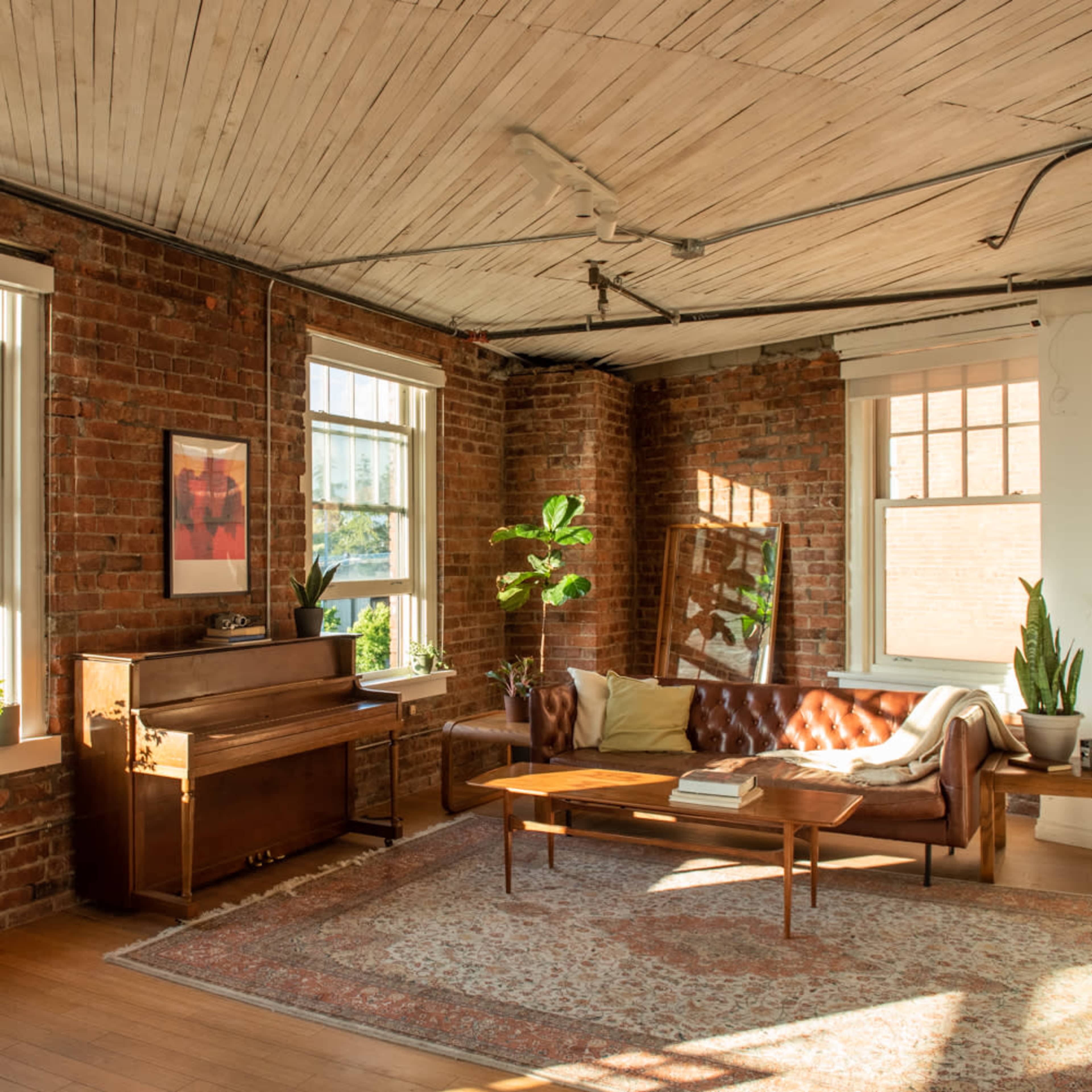 A cozy living room with exposed brick walls, large windows, and a combination of a leather sofa, a wooden piano, and a coffee table on a patterned rug.