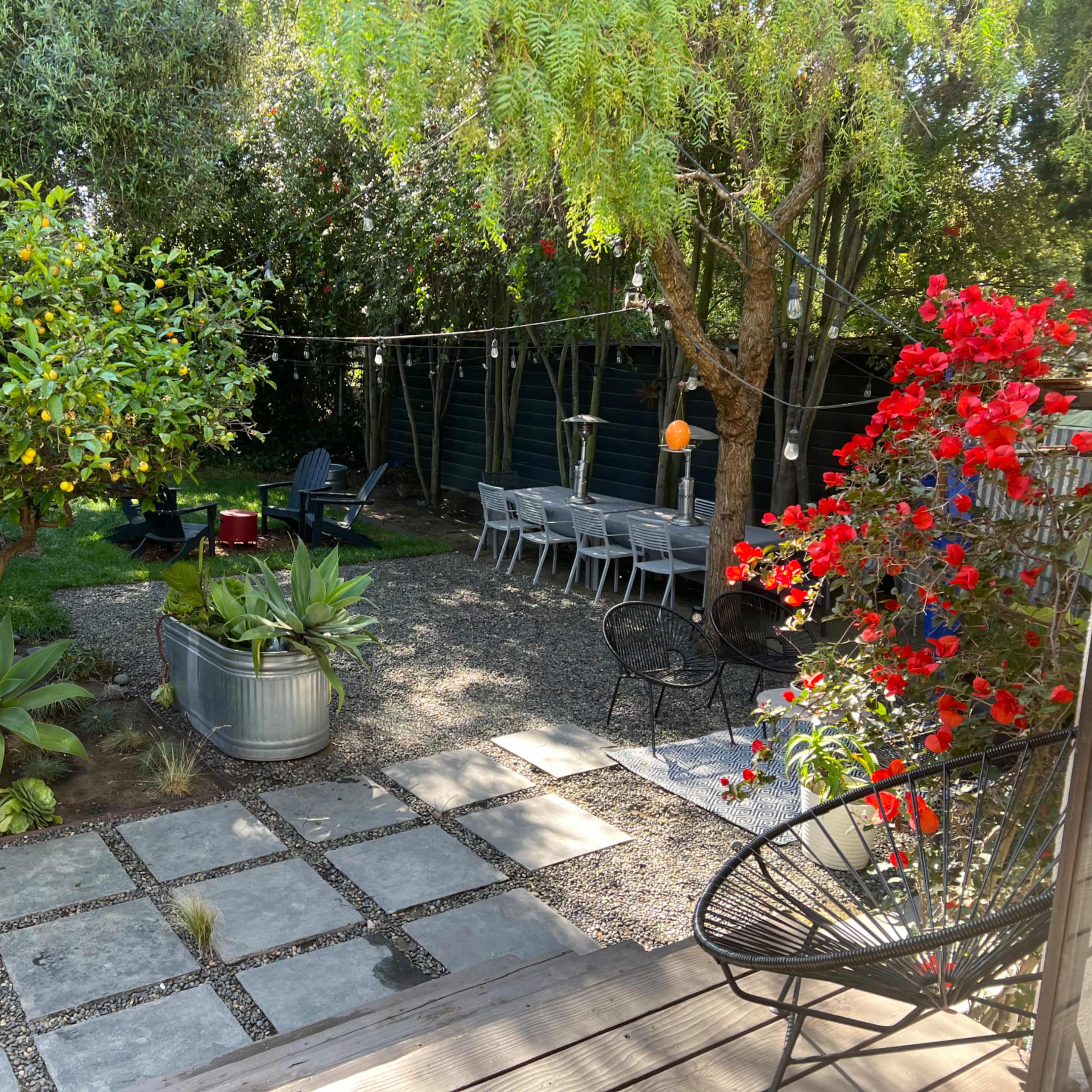 A backyard garden featuring a variety of plants, a dining table set up, and a pathway made of stone tiles.