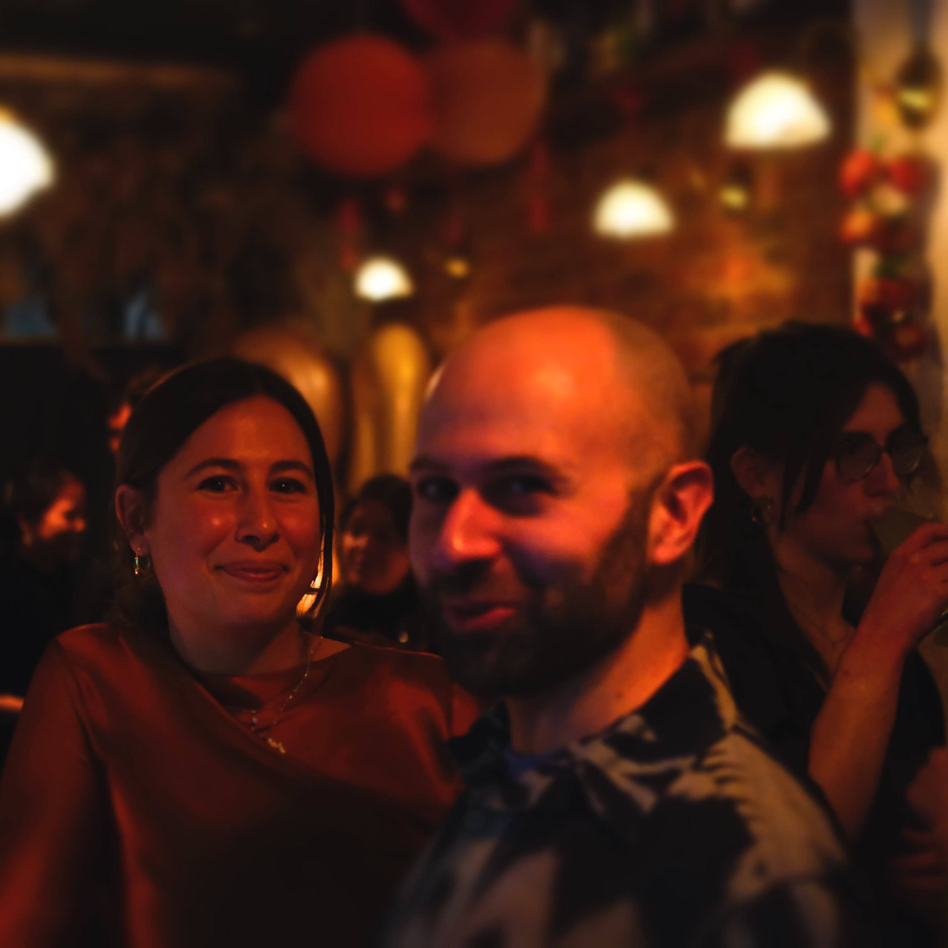 A man and a woman stand close together in a dimly lit setting with soft lighting and decorative elements in the background.