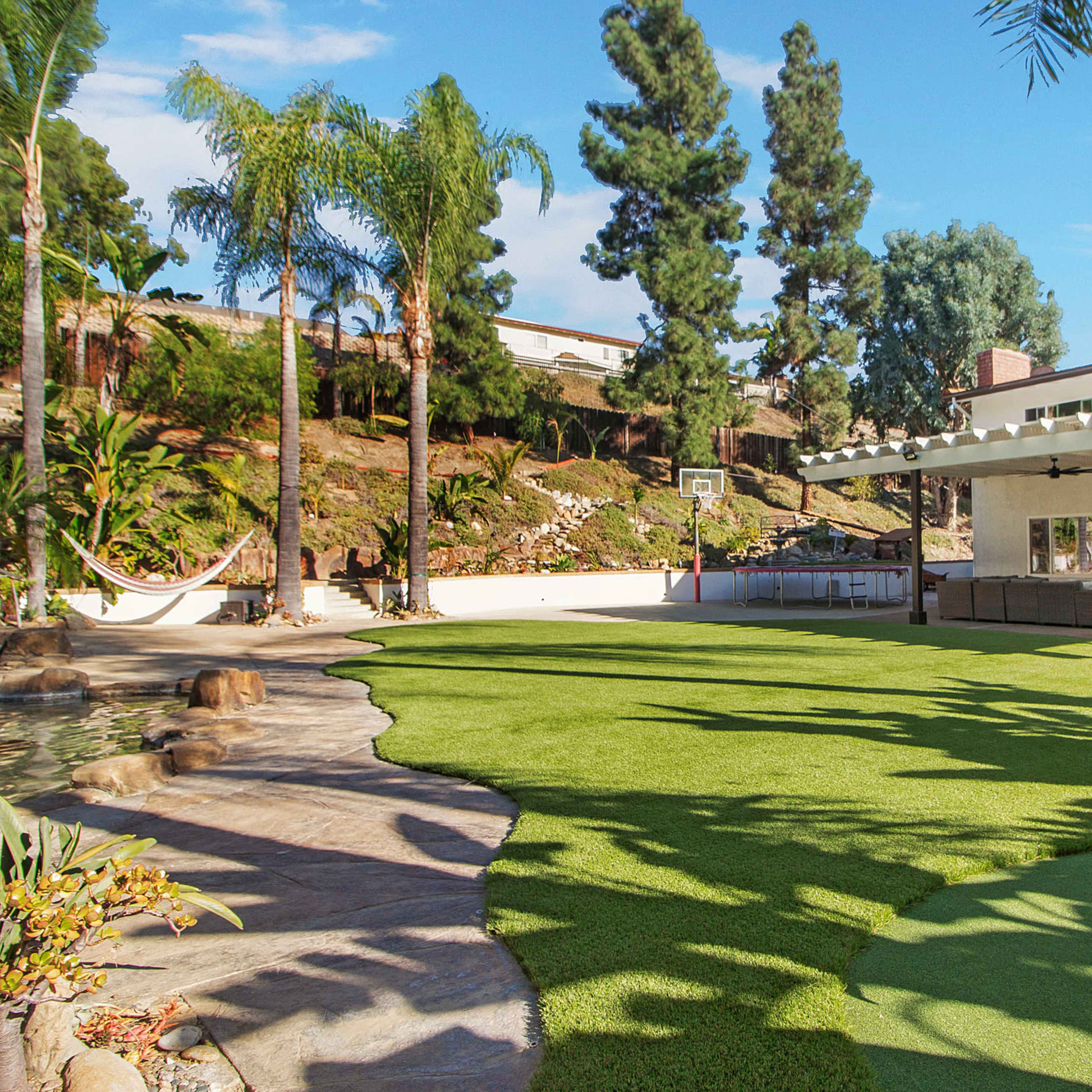 A landscaped backyard featuring a putting green, a patio with outdoor seating, palm trees, and a nearby basketball hoop.