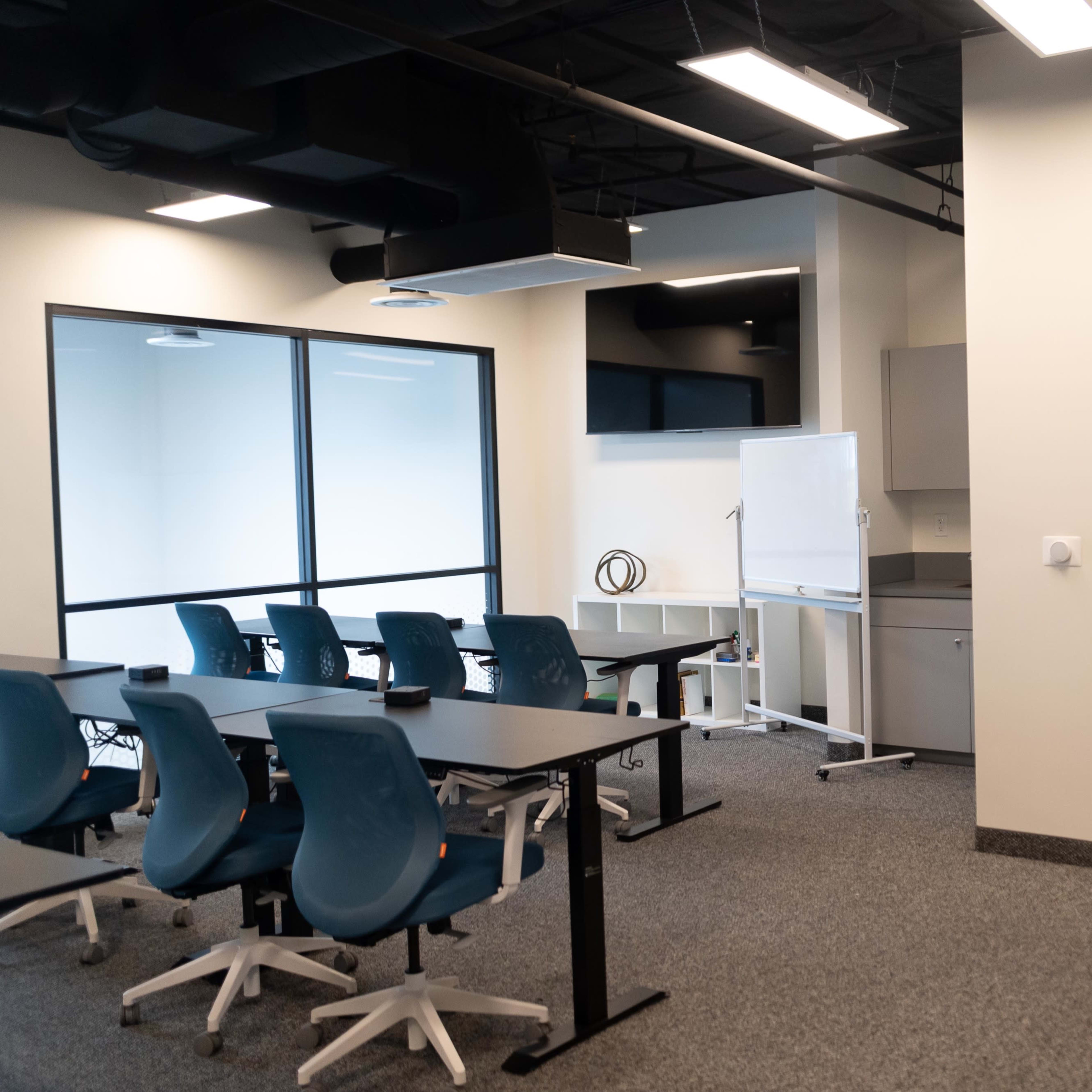 A modern office space with a conference table, chairs, and a small kitchenette, alongside a restroom door.