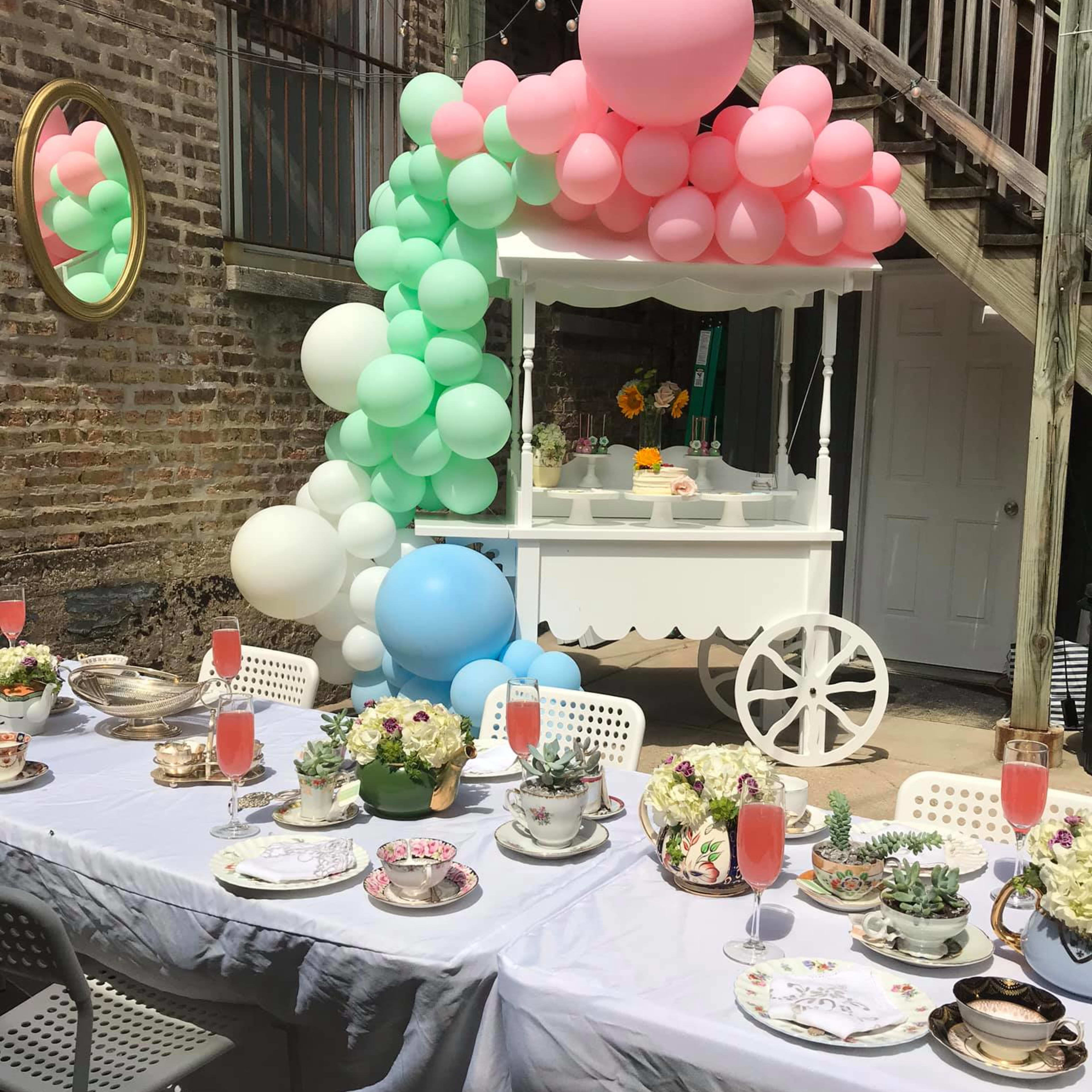 A table is set for a celebration with decorative tableware and pink drinks, alongside a balloon arch and a food cart in an outdoor space.