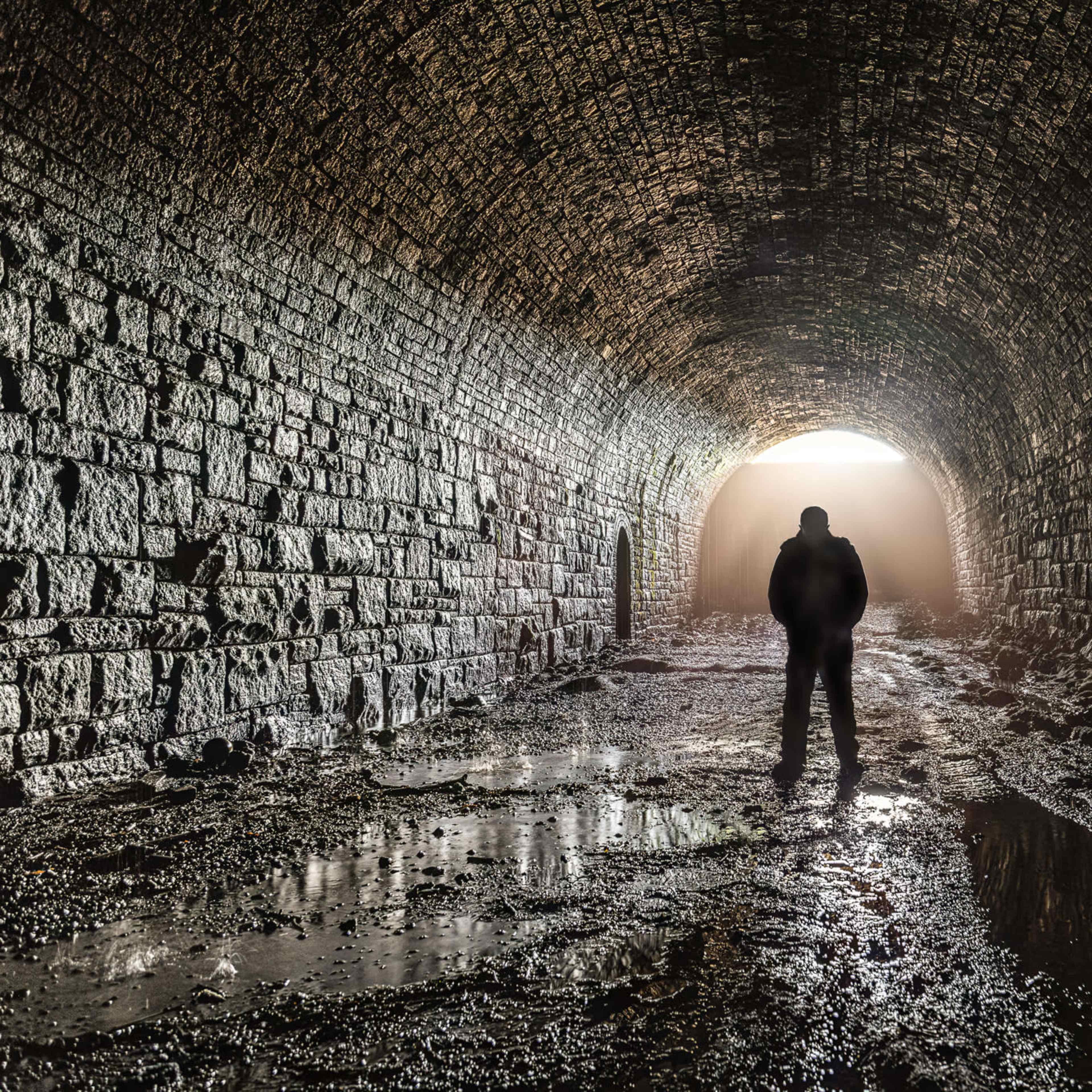 A person stands in a dimly lit stone tunnel, with light streaming in from the entrance and water pooling on the ground.