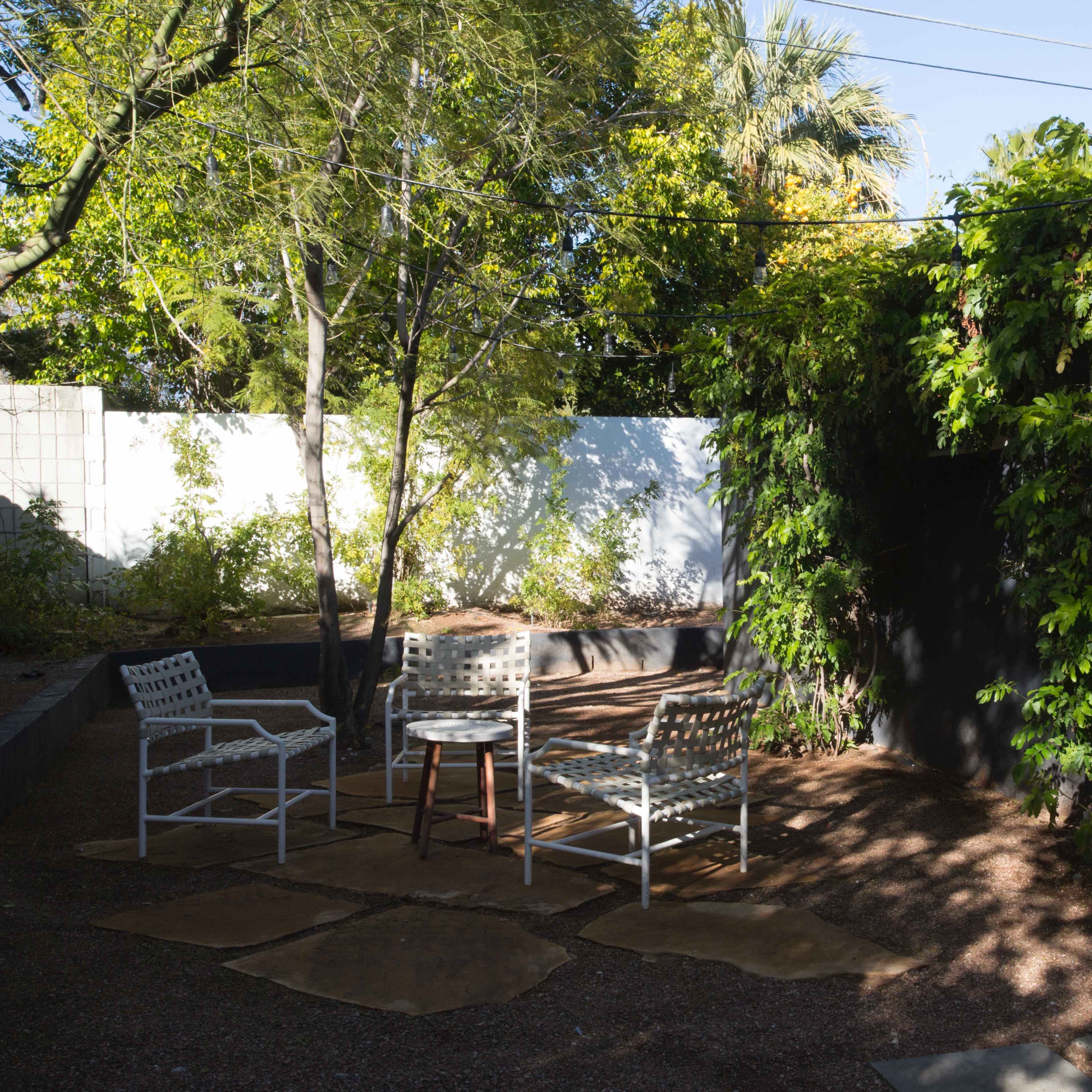 A small outdoor seating area features three white chairs and a round table surrounded by greenery and a wall.