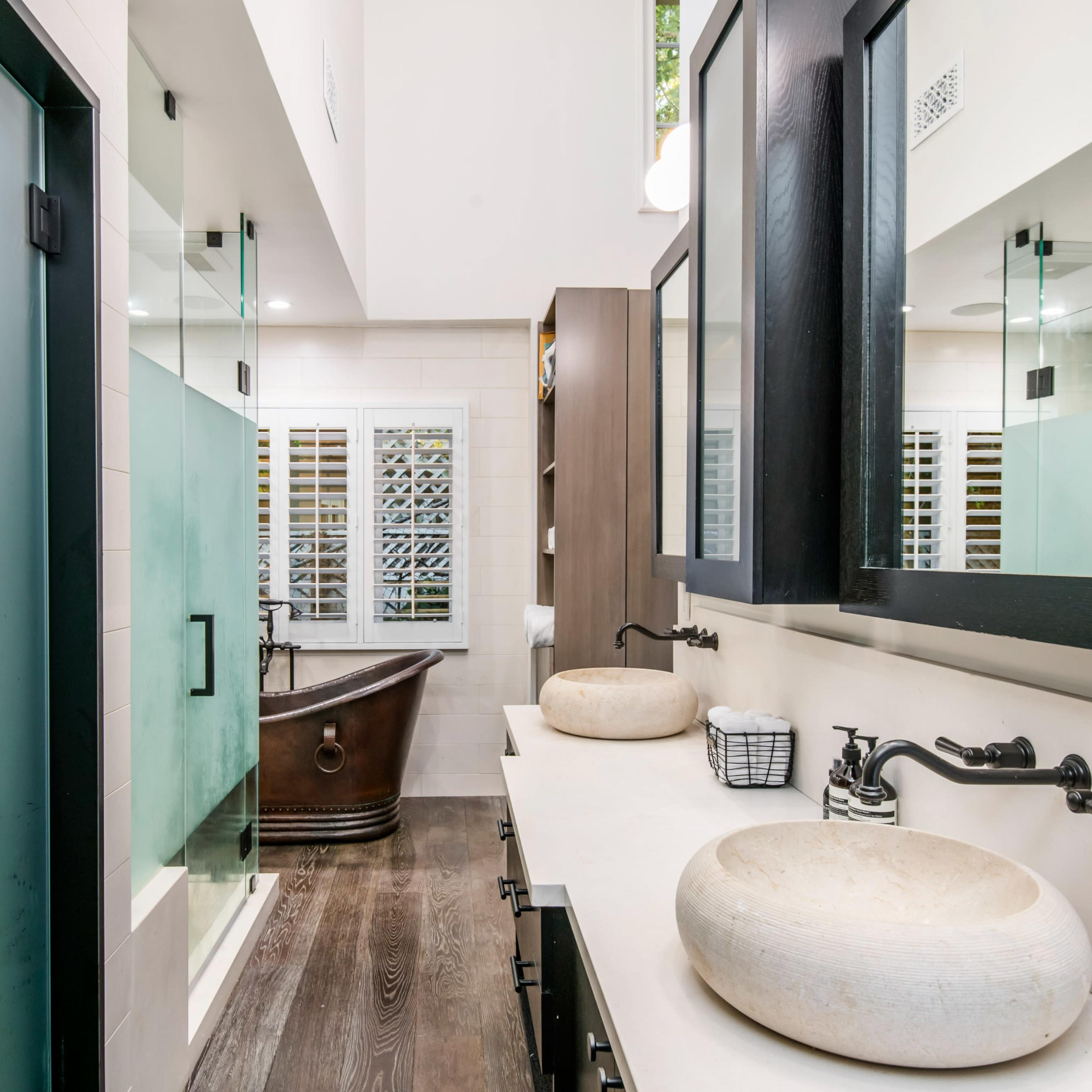 A modern bathroom featuring a double vanity with stone sinks, a freestanding bathtub, and glass shower doors.