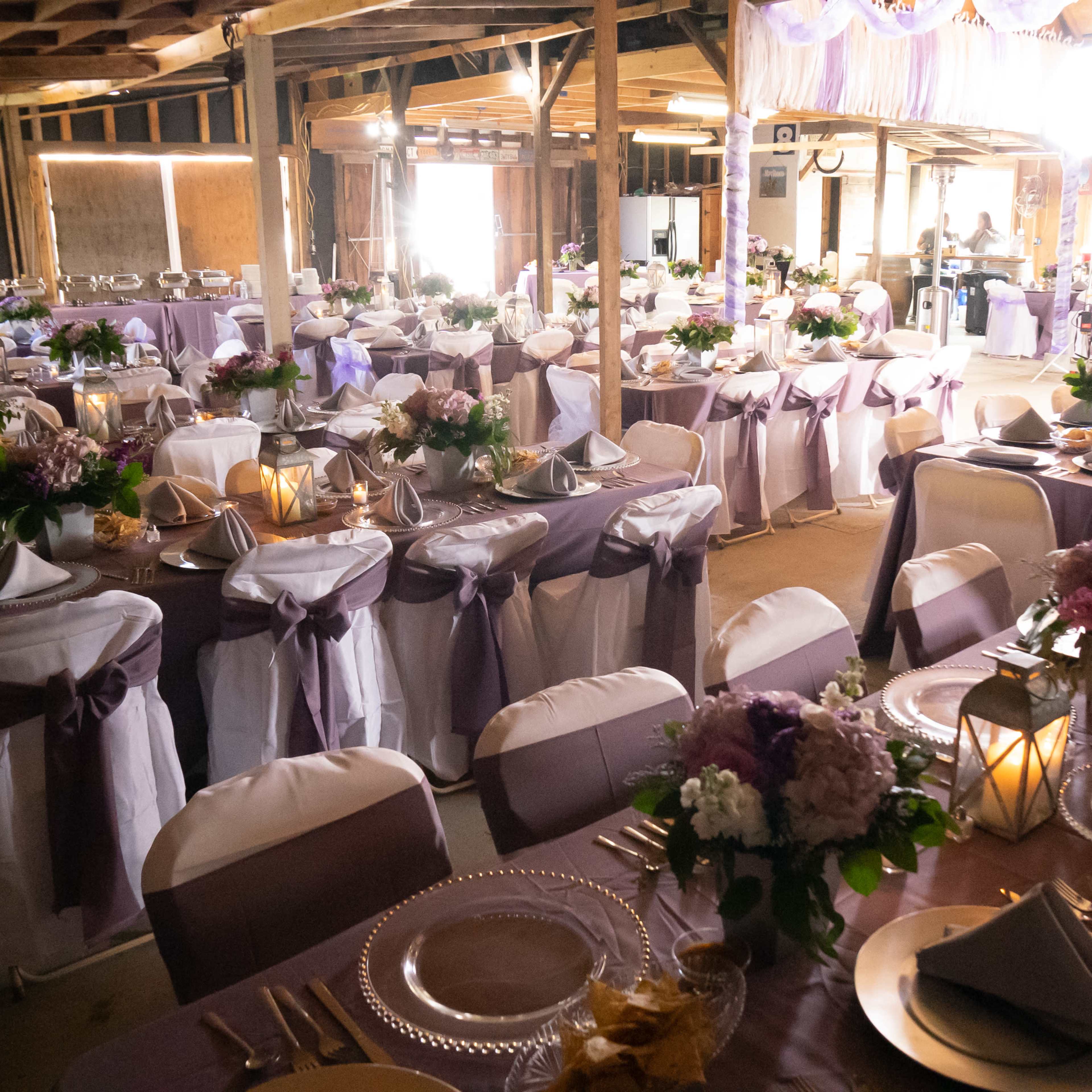 A spacious banquet hall is set up with elegantly arranged tables adorned with purple and white tablecloths, flowers, and place settings.