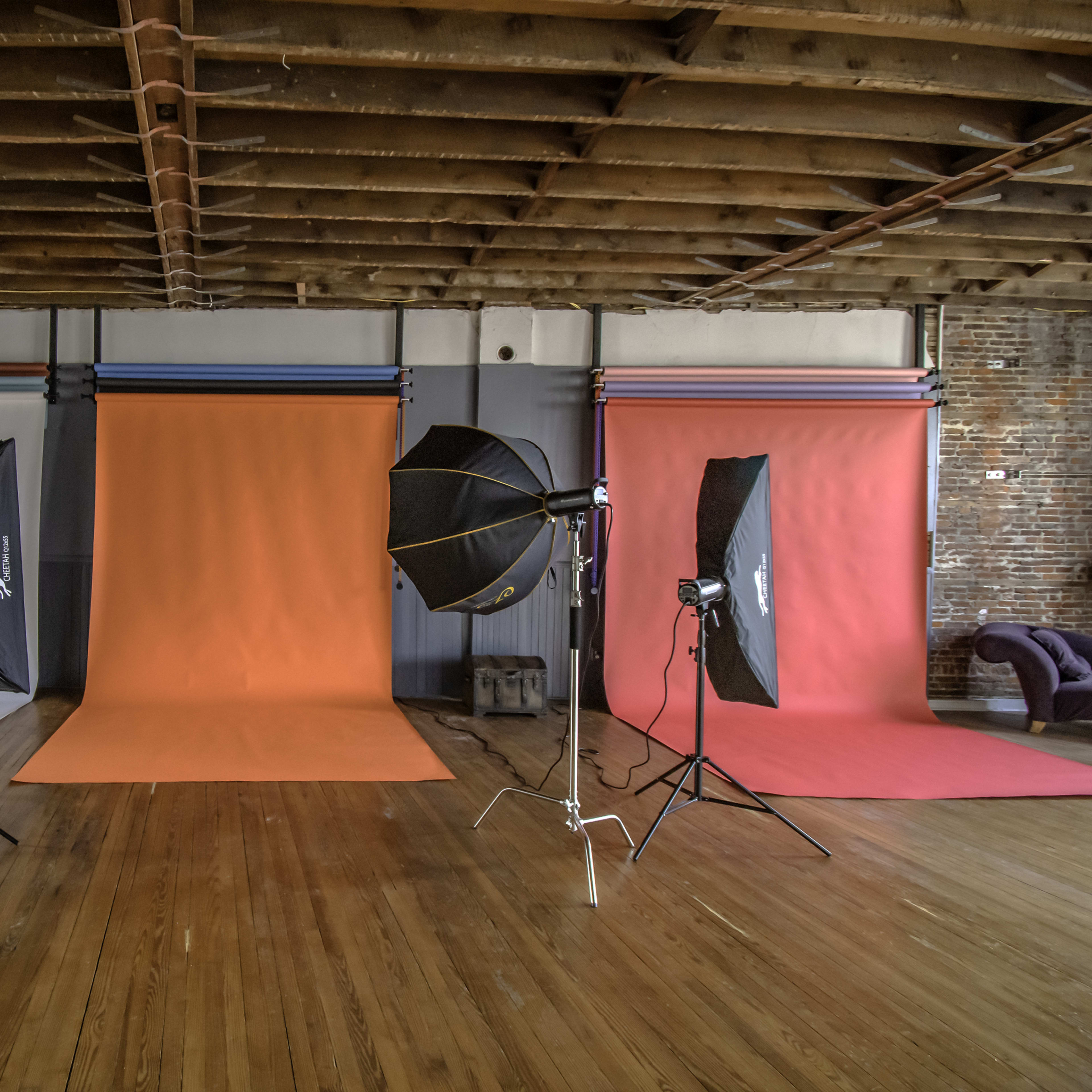 A photography studio with three backdrop setups, including a gray, orange, and light blue background, along with multiple lighting equipment positioned around the space.
