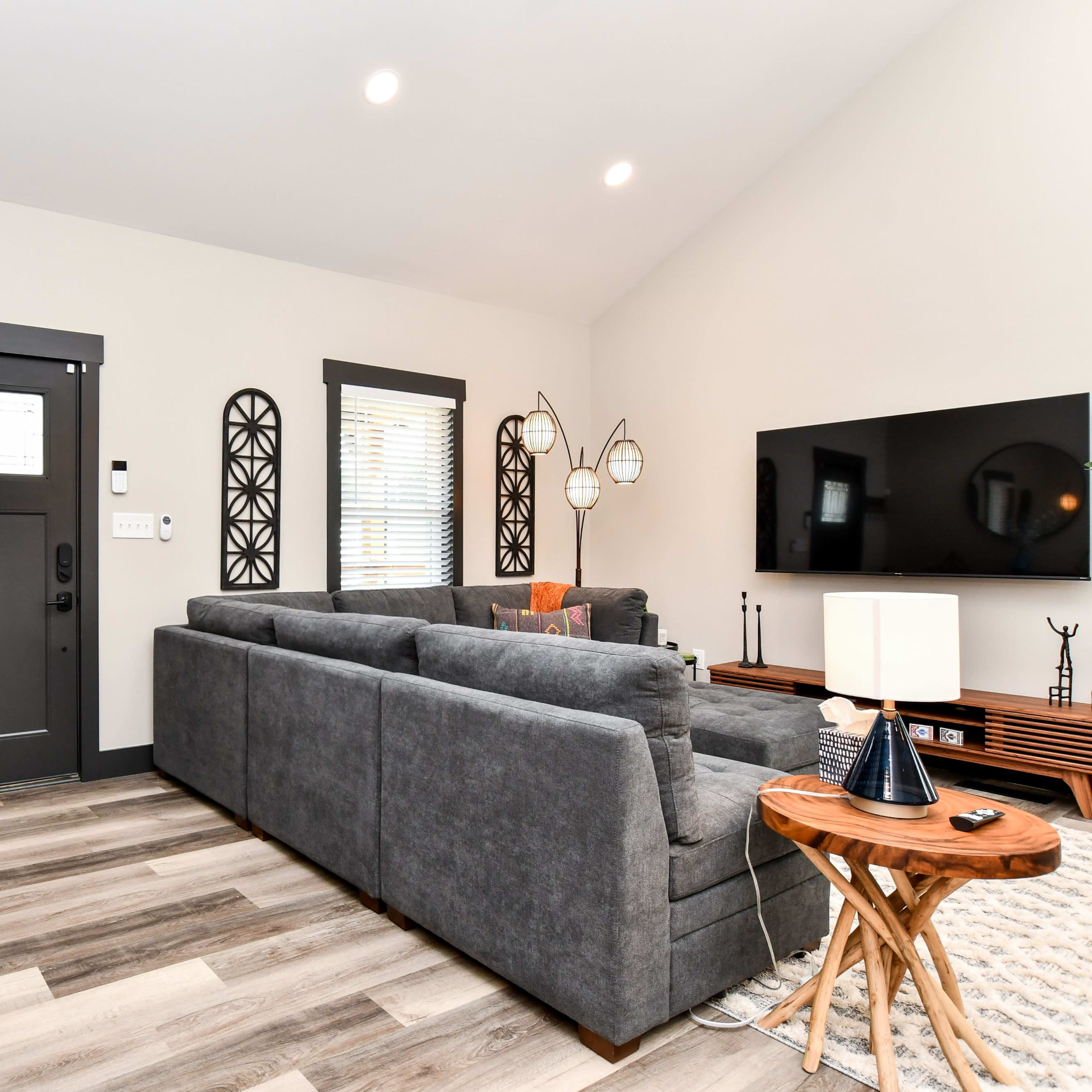 A modern living room with a gray sectional sofa, a wooden console table, a circular mirror, and a flat-screen TV mounted on the wall.
