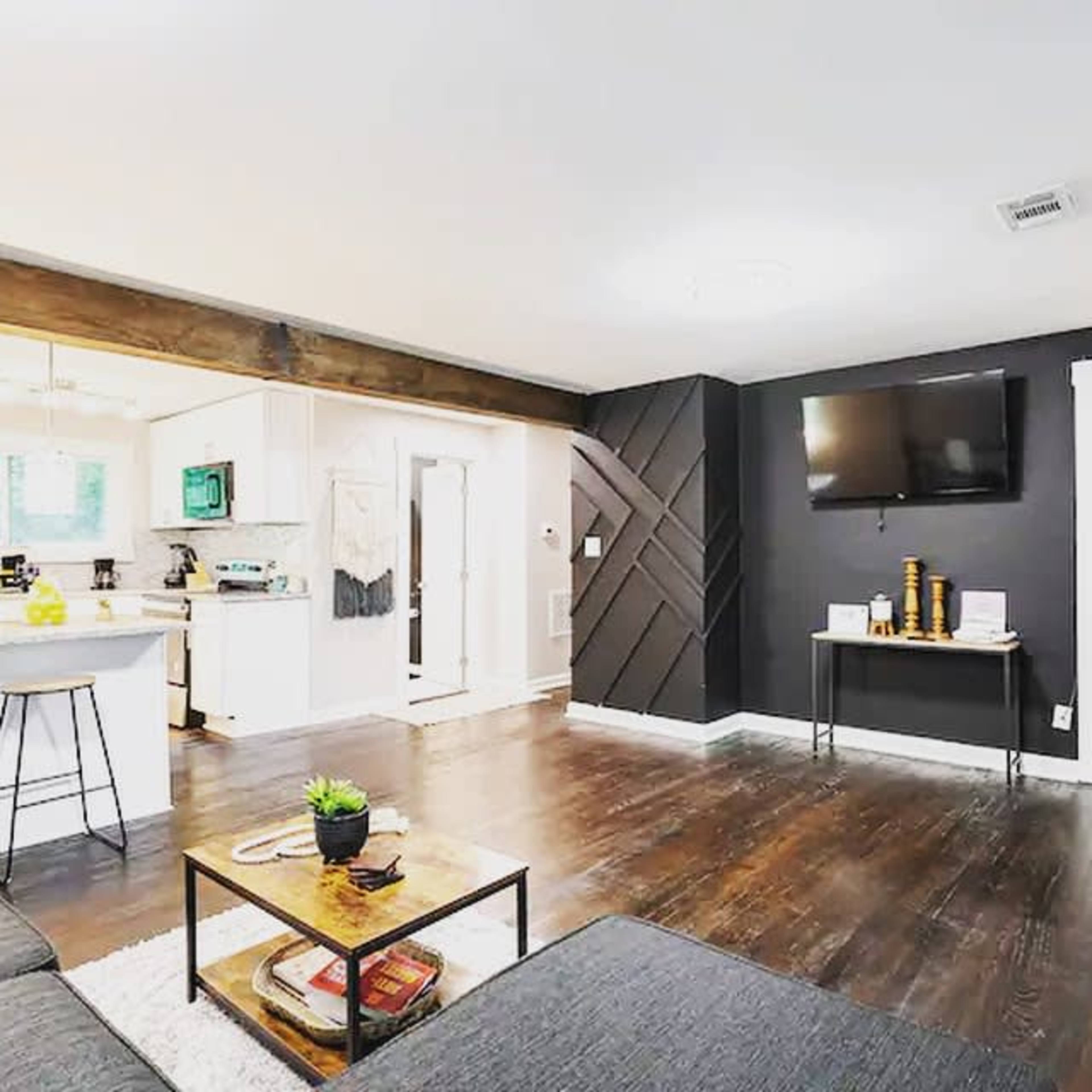 A living room features a dark accent wall with a mounted television, a sofa, a coffee table, and a view into a kitchen area.