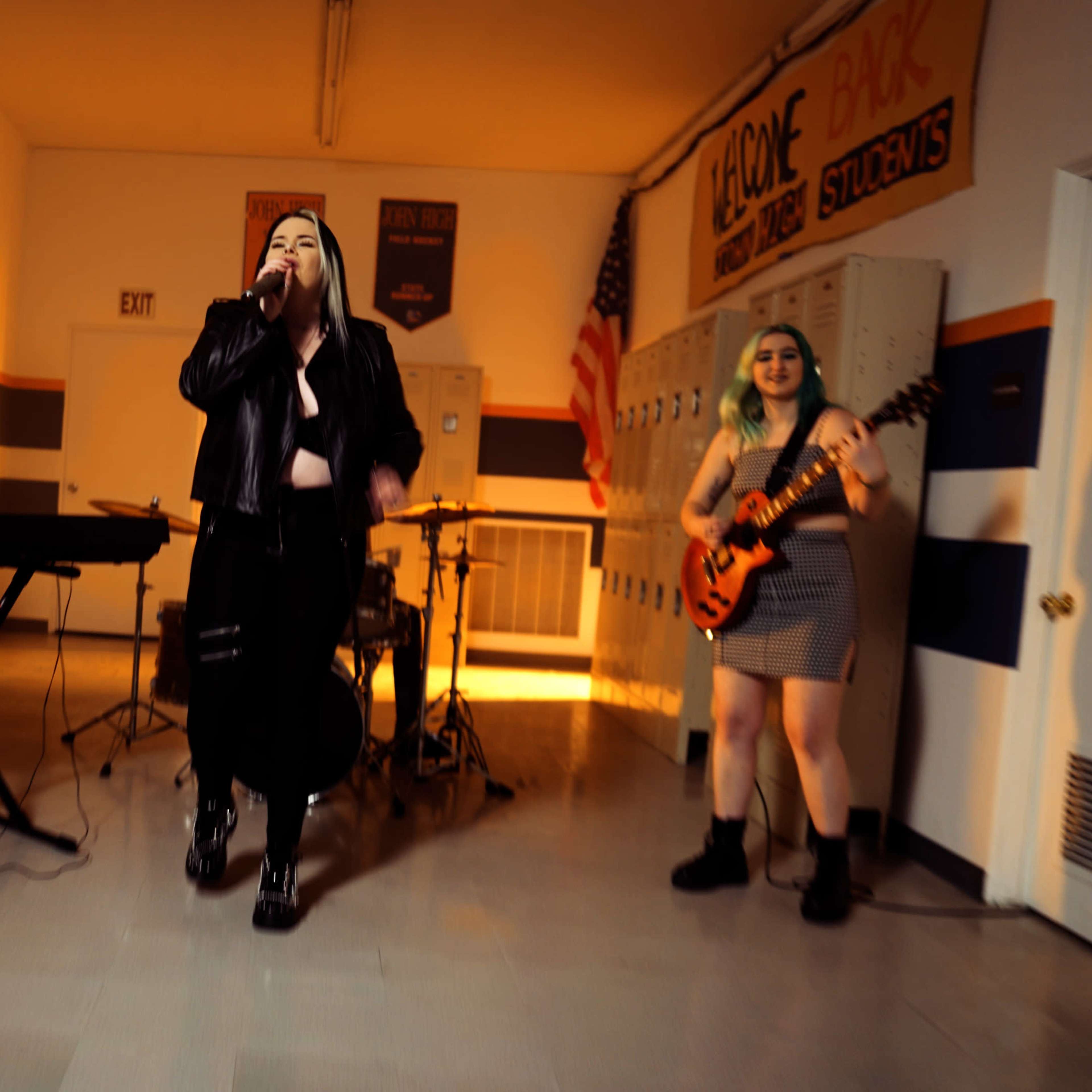 A band performs in a school hallway with lockers in the background, featuring a singer, keyboardist, and guitarist.
