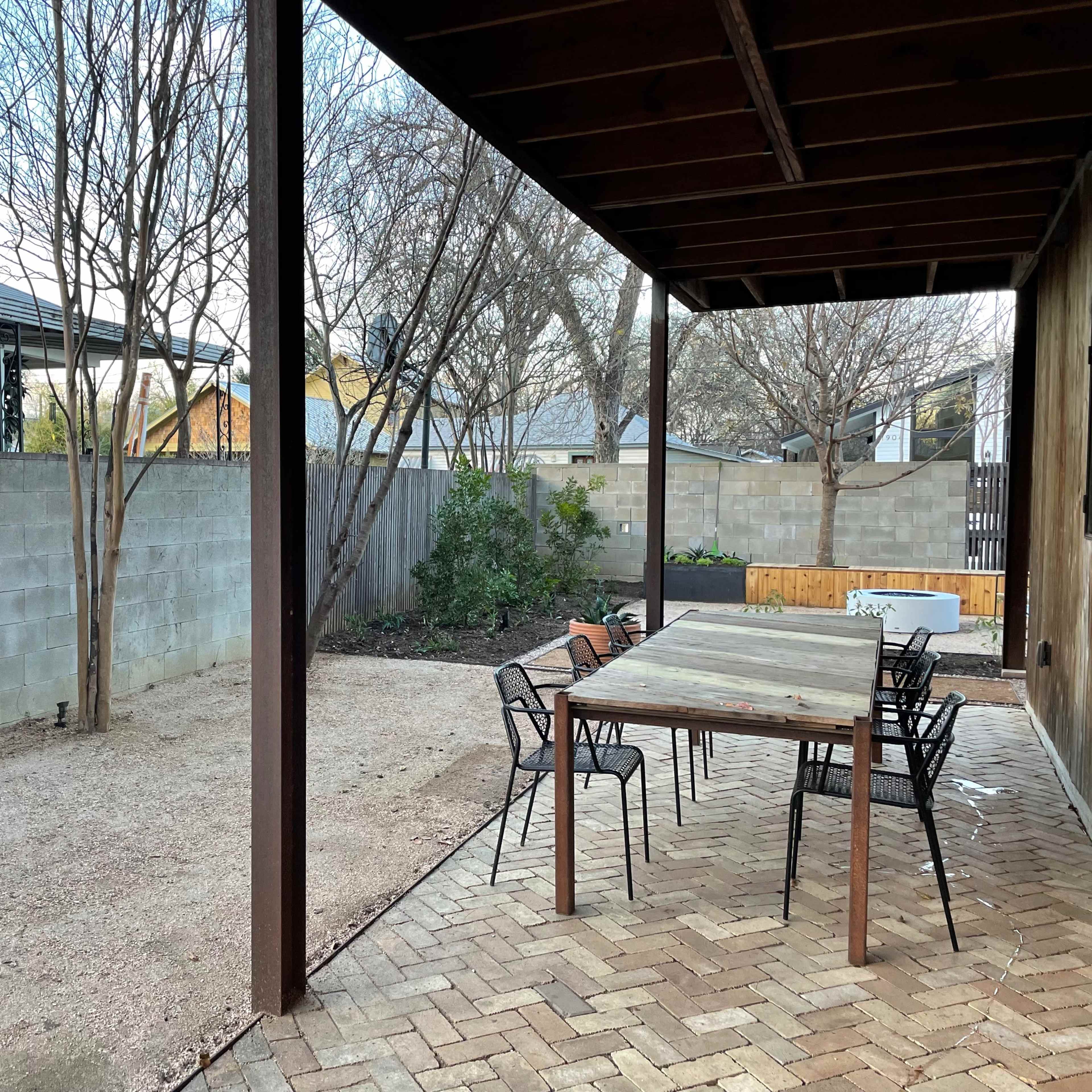A large wooden table with chairs sits on a brick patio under a shaded roof, adjacent to a gravel area and surrounded by a low stone wall and trees.