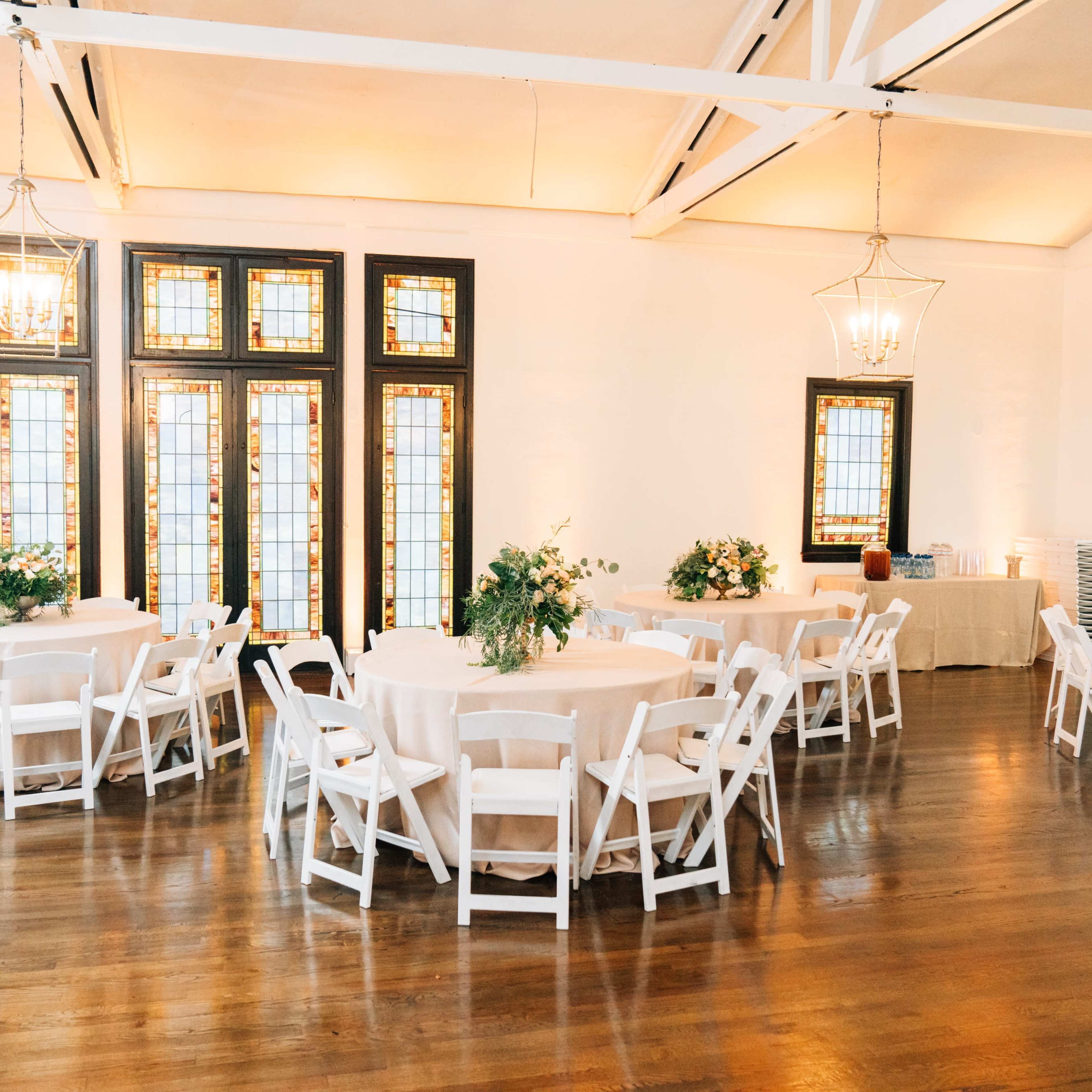 A spacious event room with several round tables covered with white tablecloths, surrounded by white chairs and light streaming through stained glass windows.