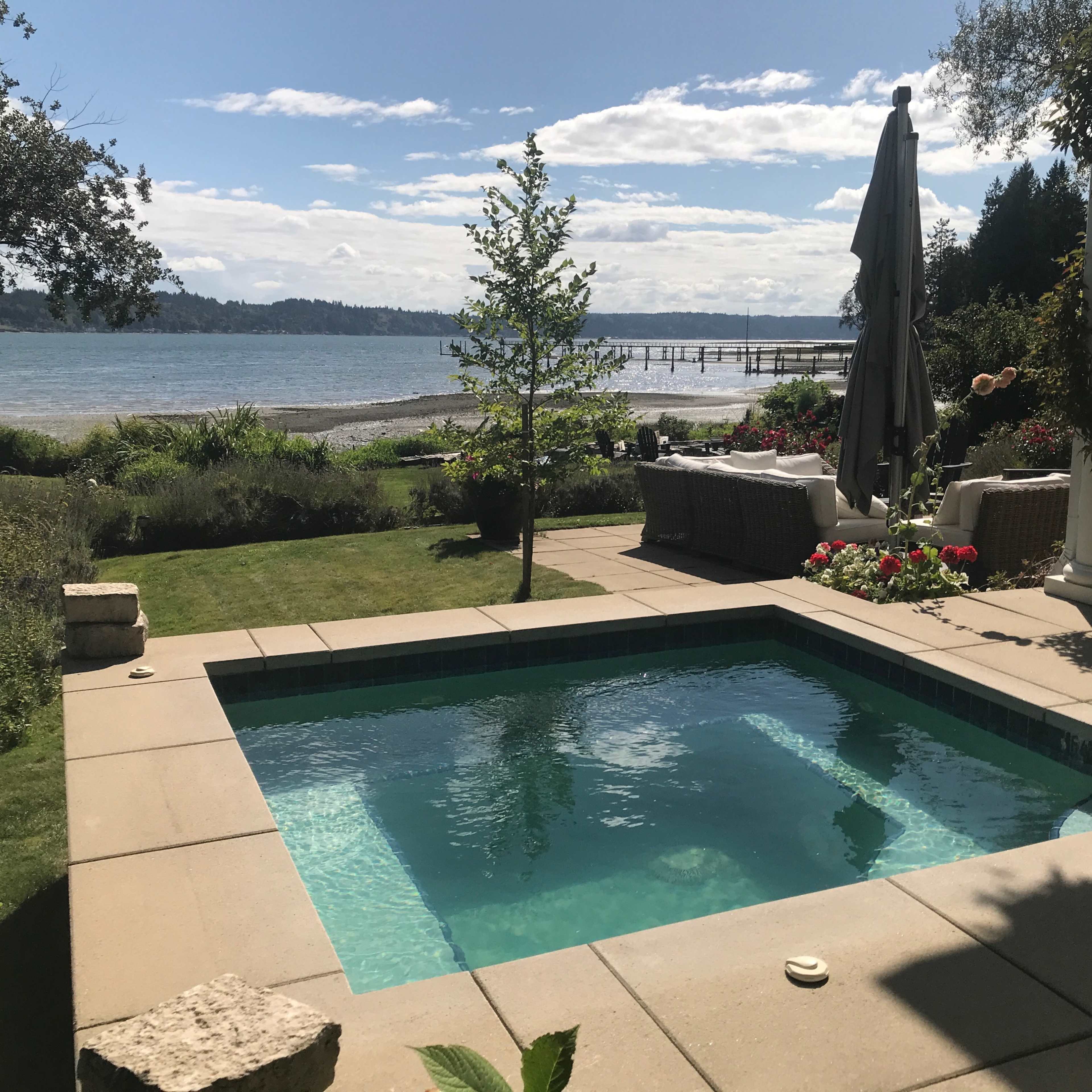A small, rectangular hot tub sits on a stone patio overlooking a body of water, surrounded by greenery and outdoor furniture.