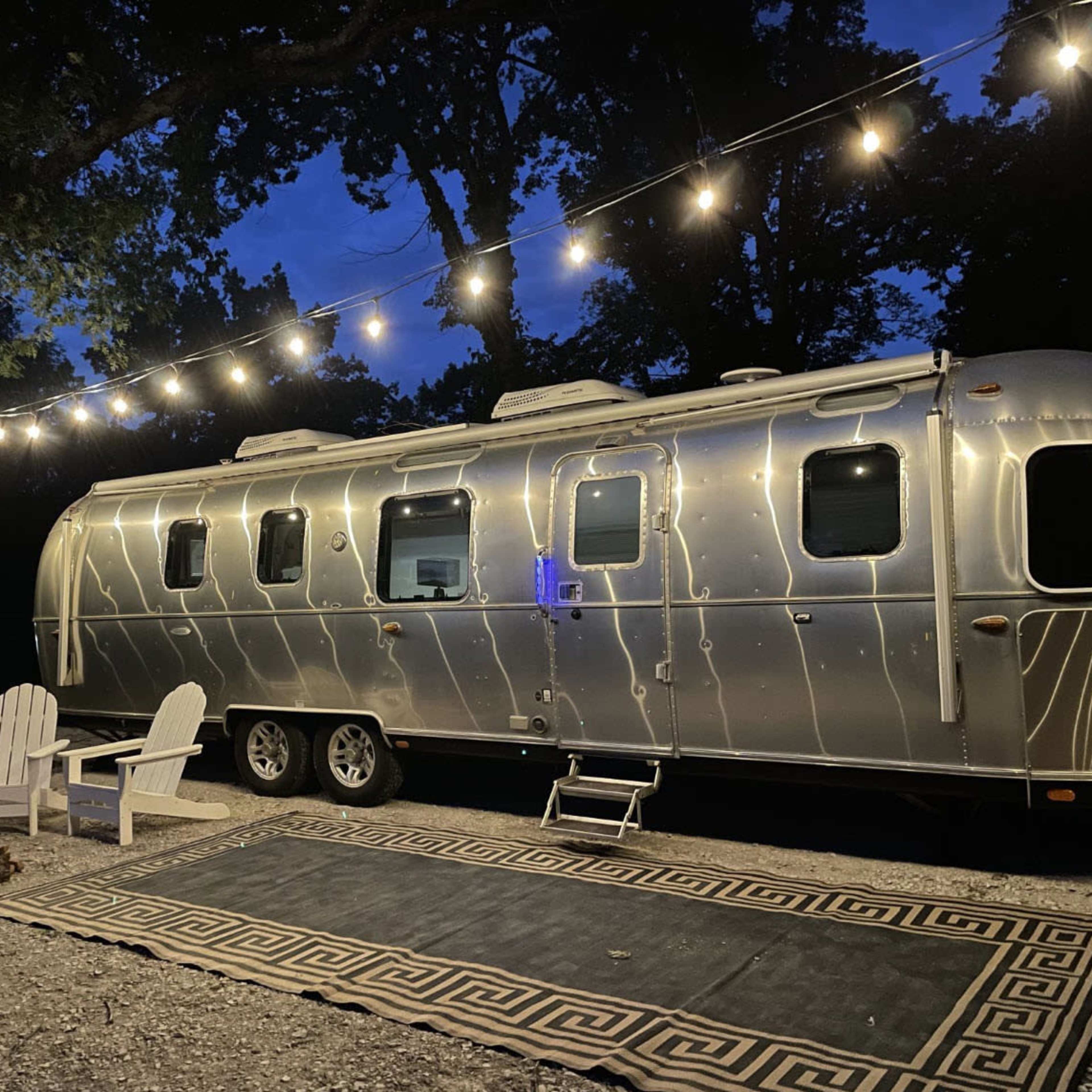 A polished silver Airstream trailer is parked under string lights next to a fire pit and two Adirondack chairs on a patterned rug.