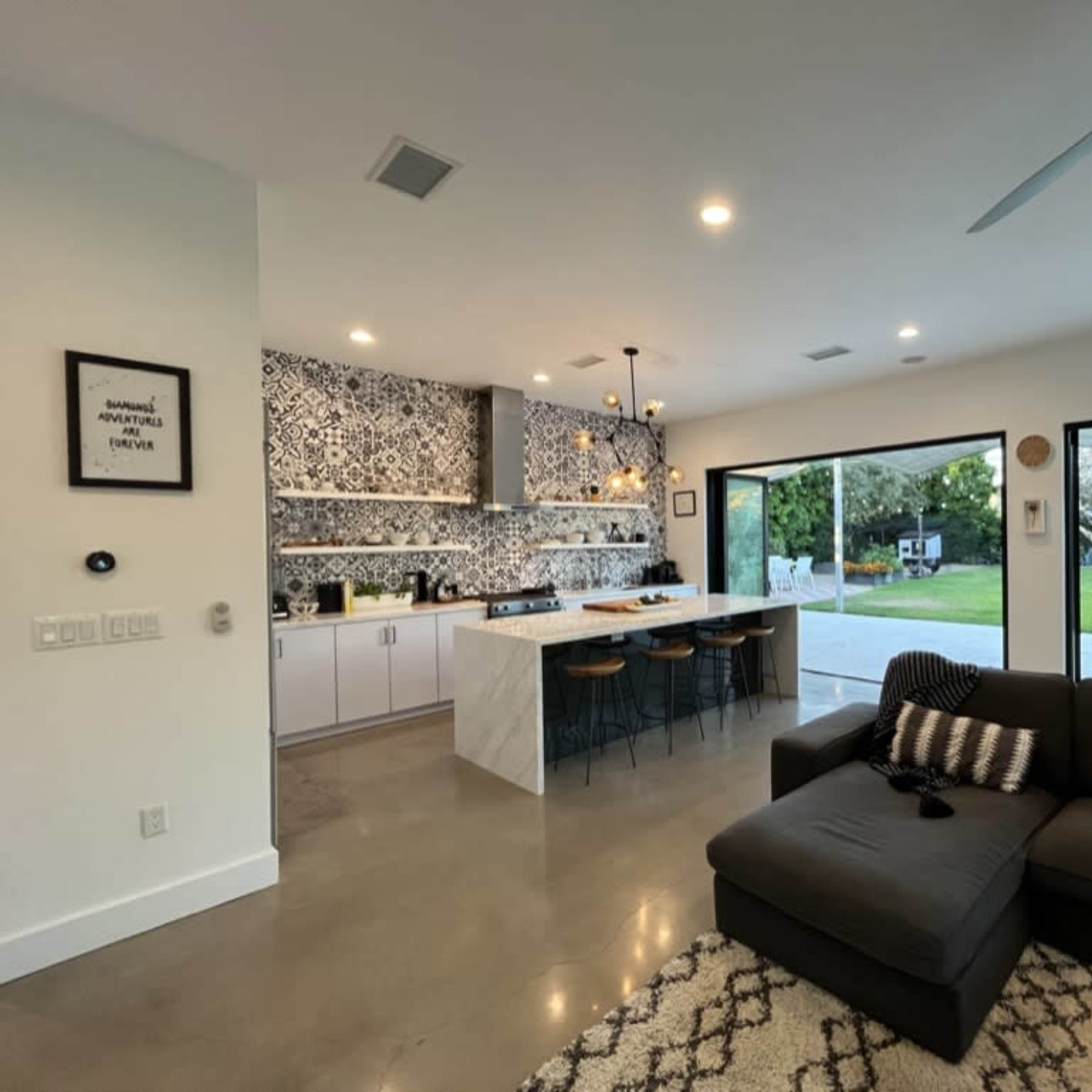 A modern kitchen and living area with a large sectional sofa, a patterned backsplash, and large windows overlooking a green outdoor space.