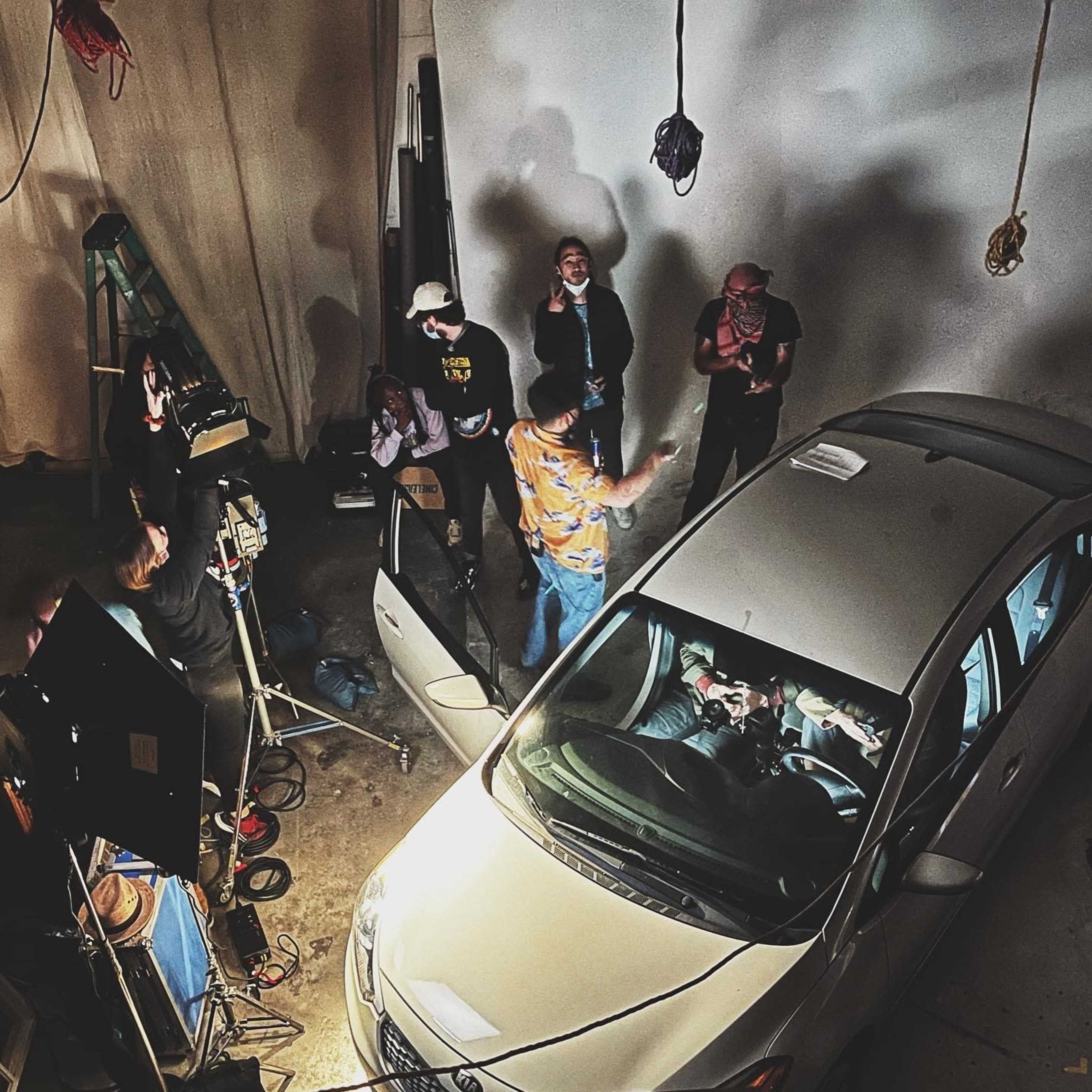 A group of people is gathered around a silver car in a dimly lit studio, with film equipment and backdrops visible in the background.