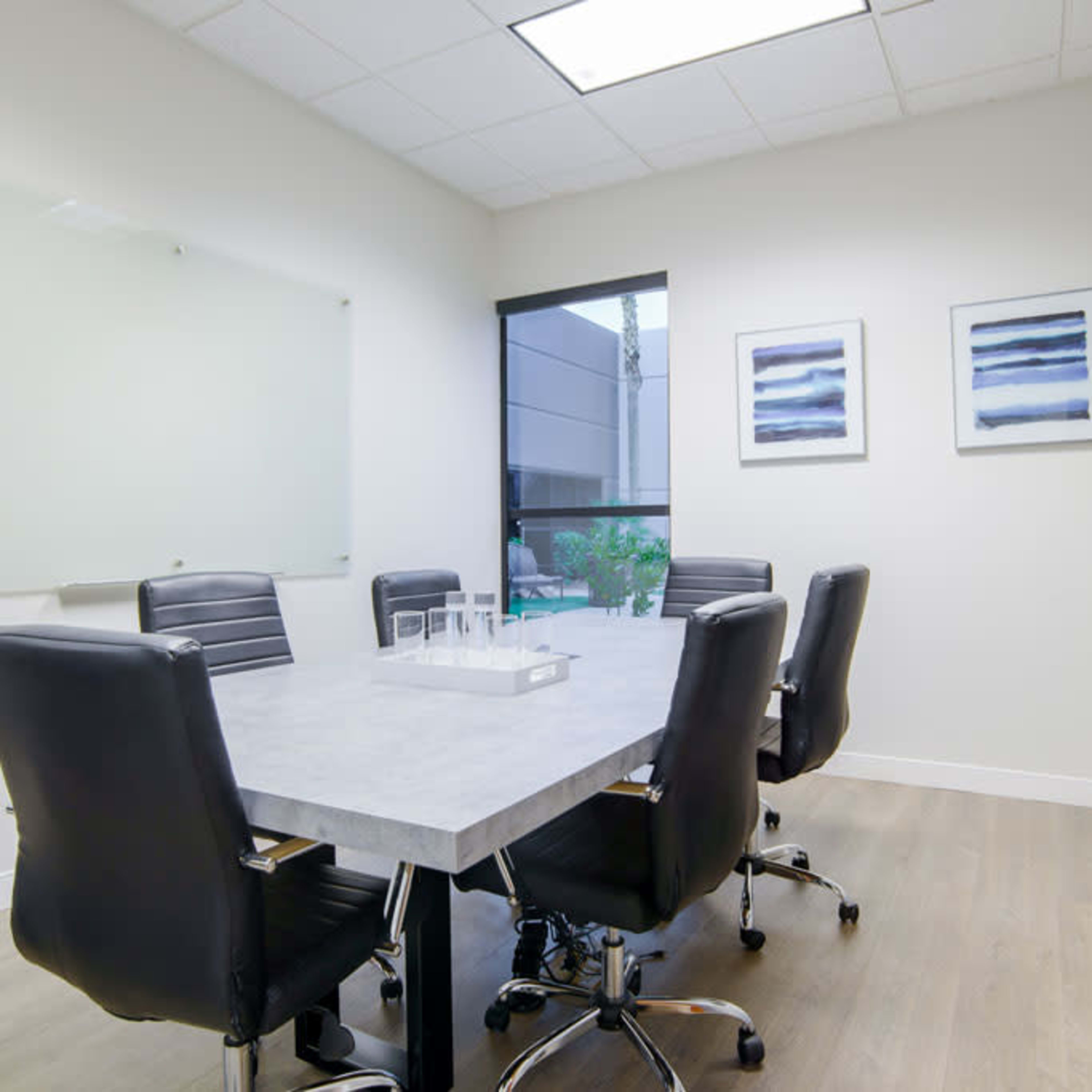 A modern conference room with a large table surrounded by black chairs, a glass whiteboard, and two framed pictures on the wall.