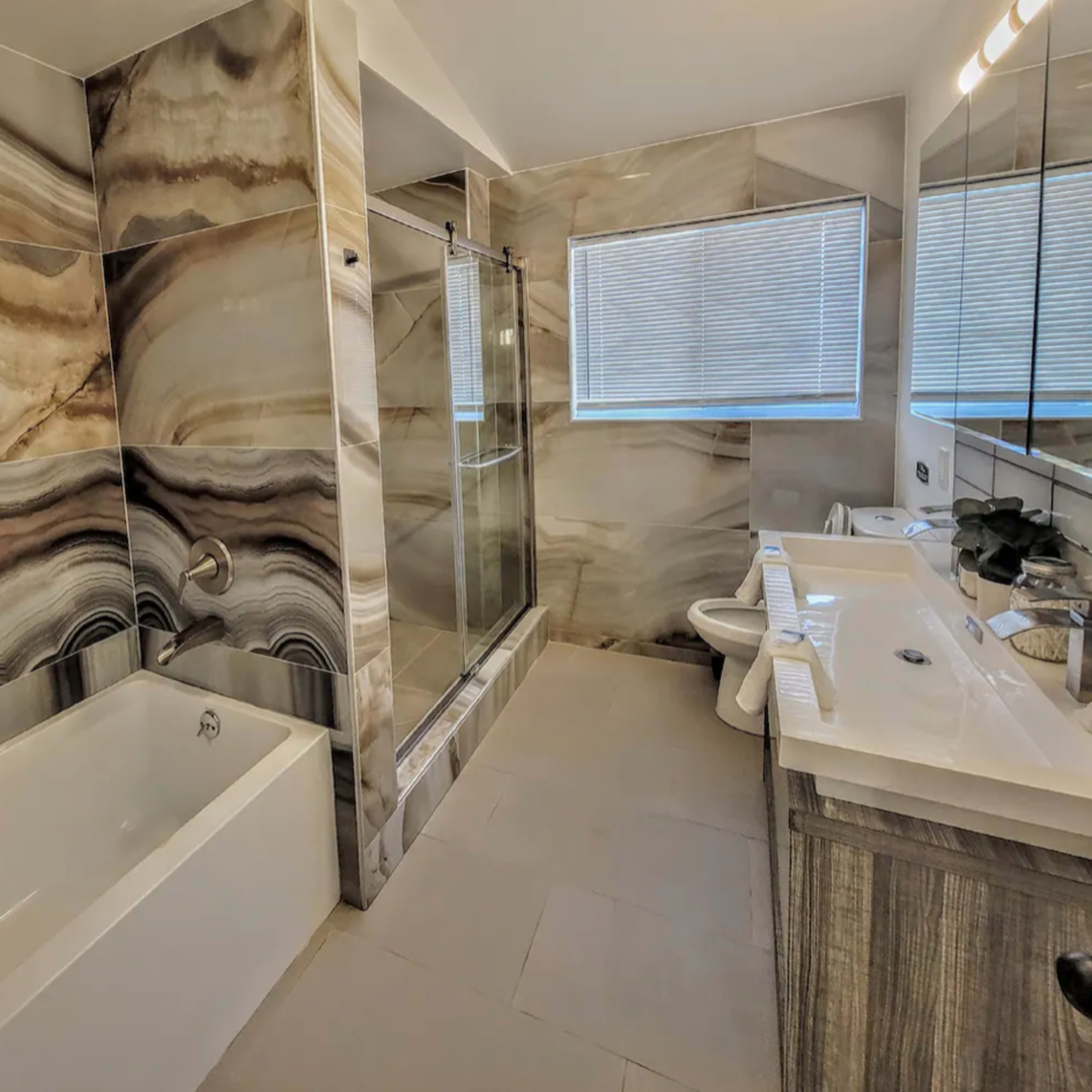 A modern bathroom featuring a bathtub, a glass shower enclosure, and dual sinks with a large mirror above them, all set against marble-patterned wall tiles.
