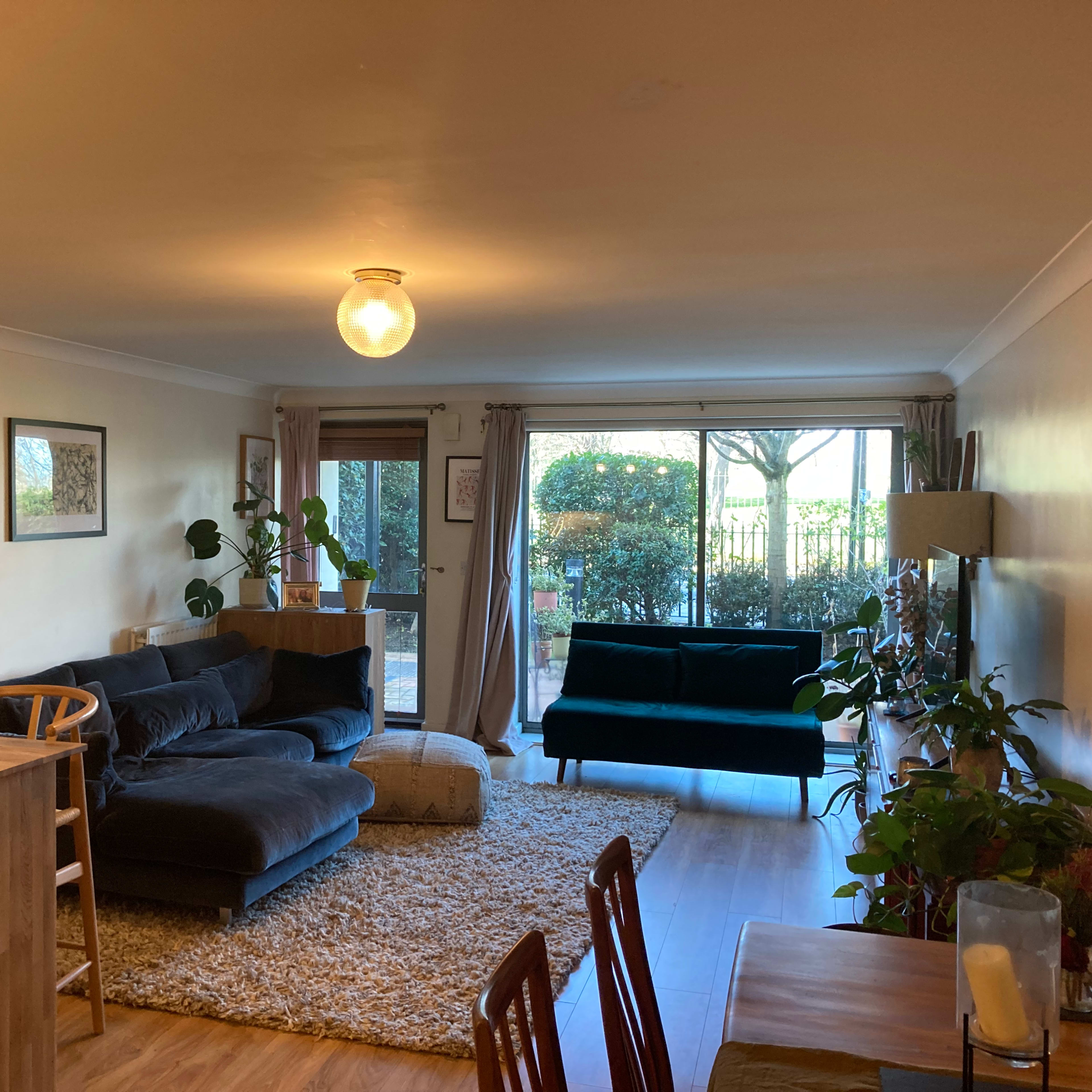 A living room features a dark sectional sofa, a green chair, a wooden dining table, and large windows with greenery visible outside.