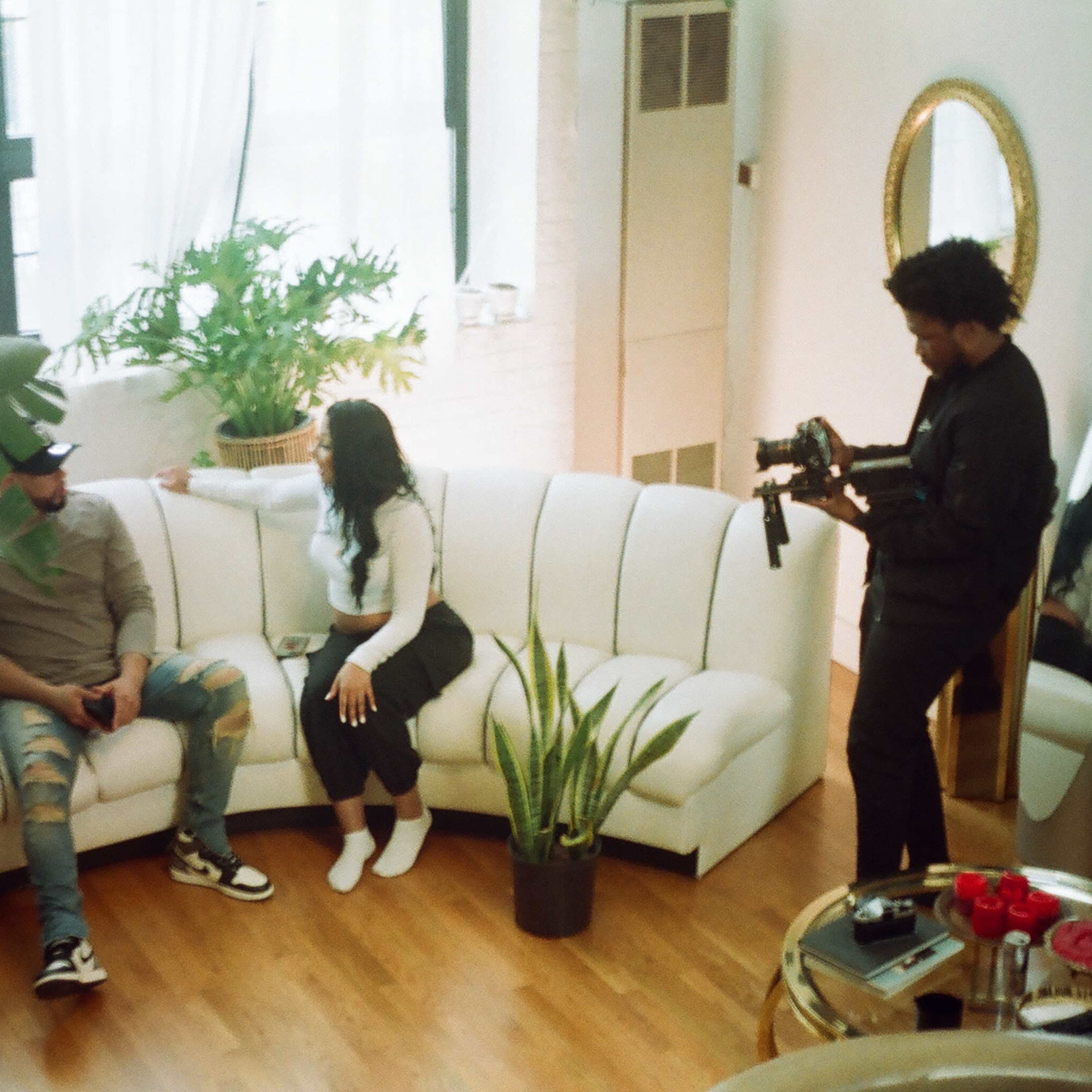 Three people are seated and conversing on a white sectional sofa in a well-lit room, surrounded by plants and a large mirror.