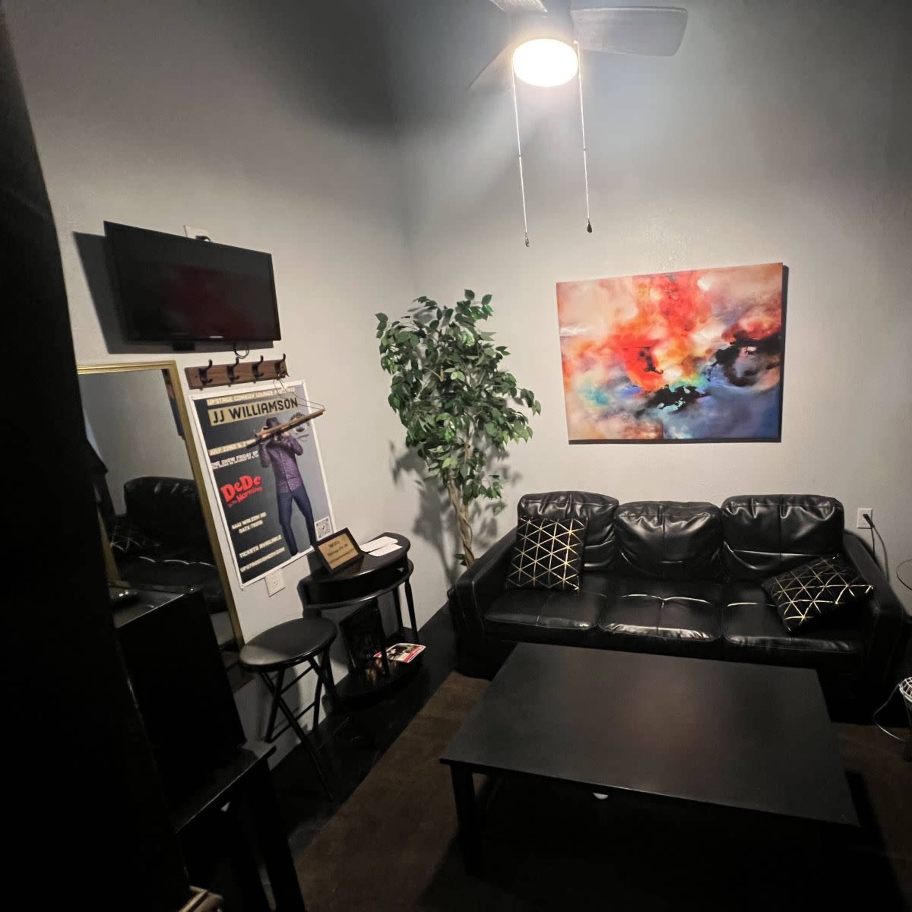 A small room featuring a black leather couch, a coffee table, a decorative plant, a wall-mounted TV, and an abstract painting.