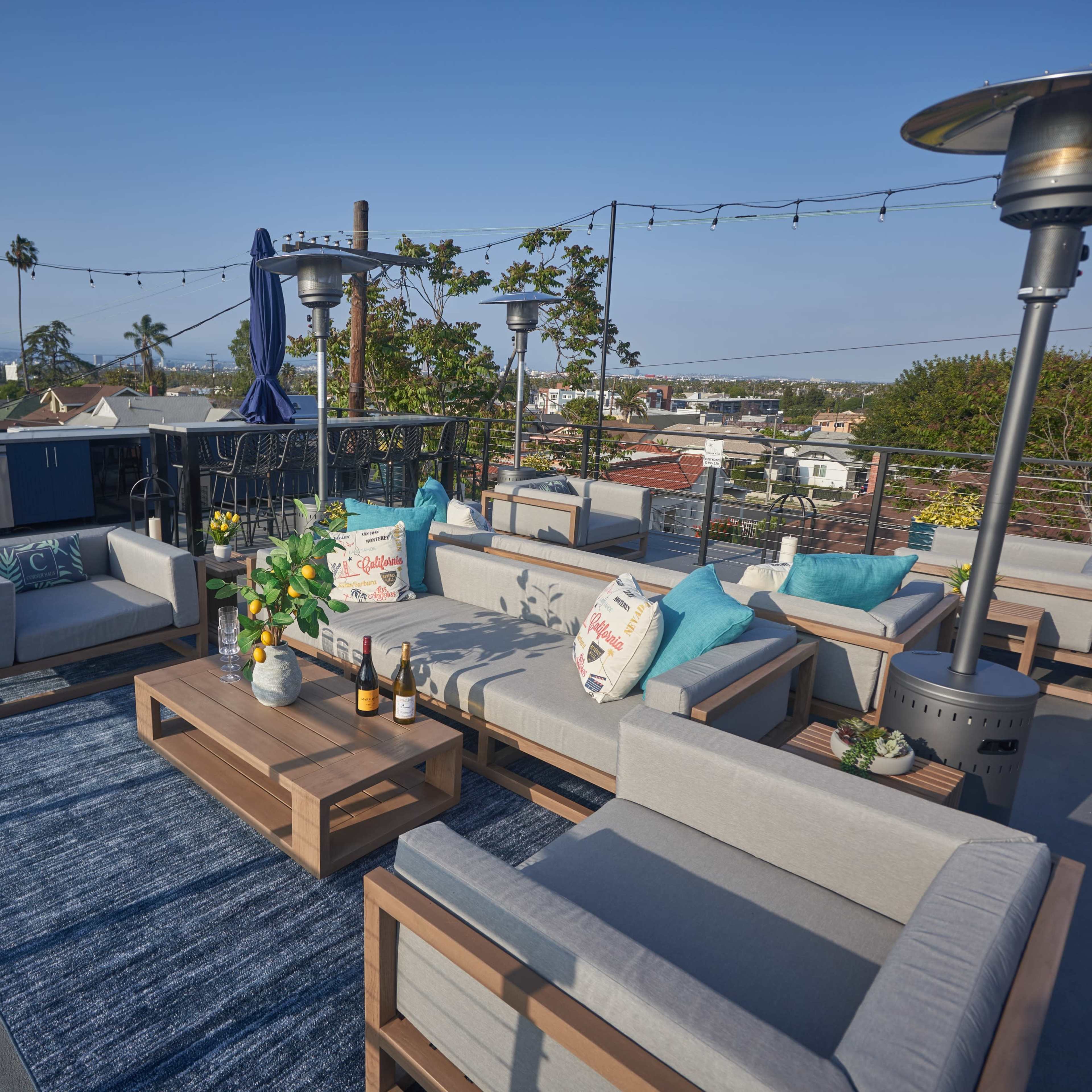 The rooftop deck features modern seating, a table with drinks, and a view of surrounding houses and palm trees.