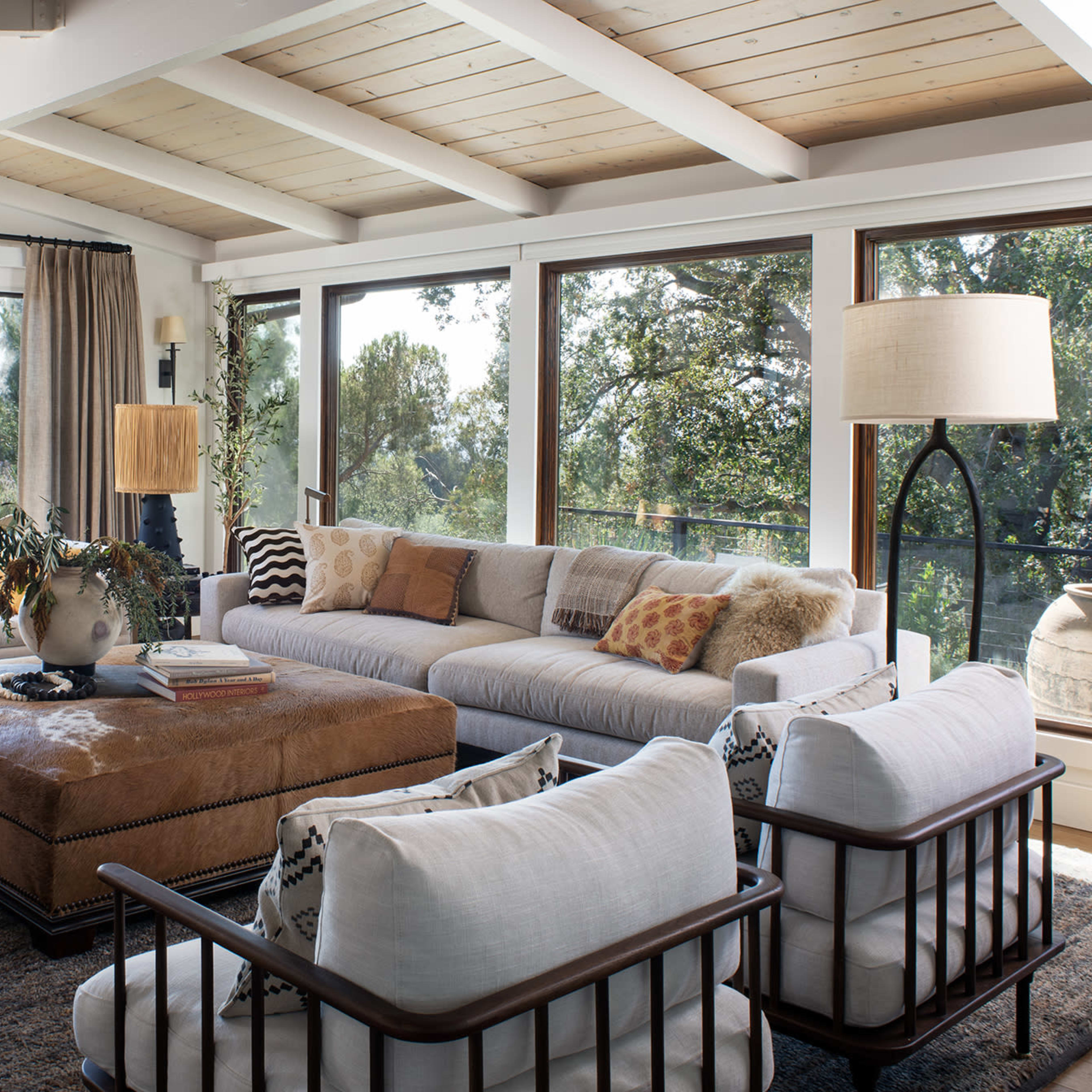 A spacious living room with large windows, showcasing a view of greenery and mountains, furnished with a sectional sofa, patterned pillows, an ottoman, and various decorative elements.