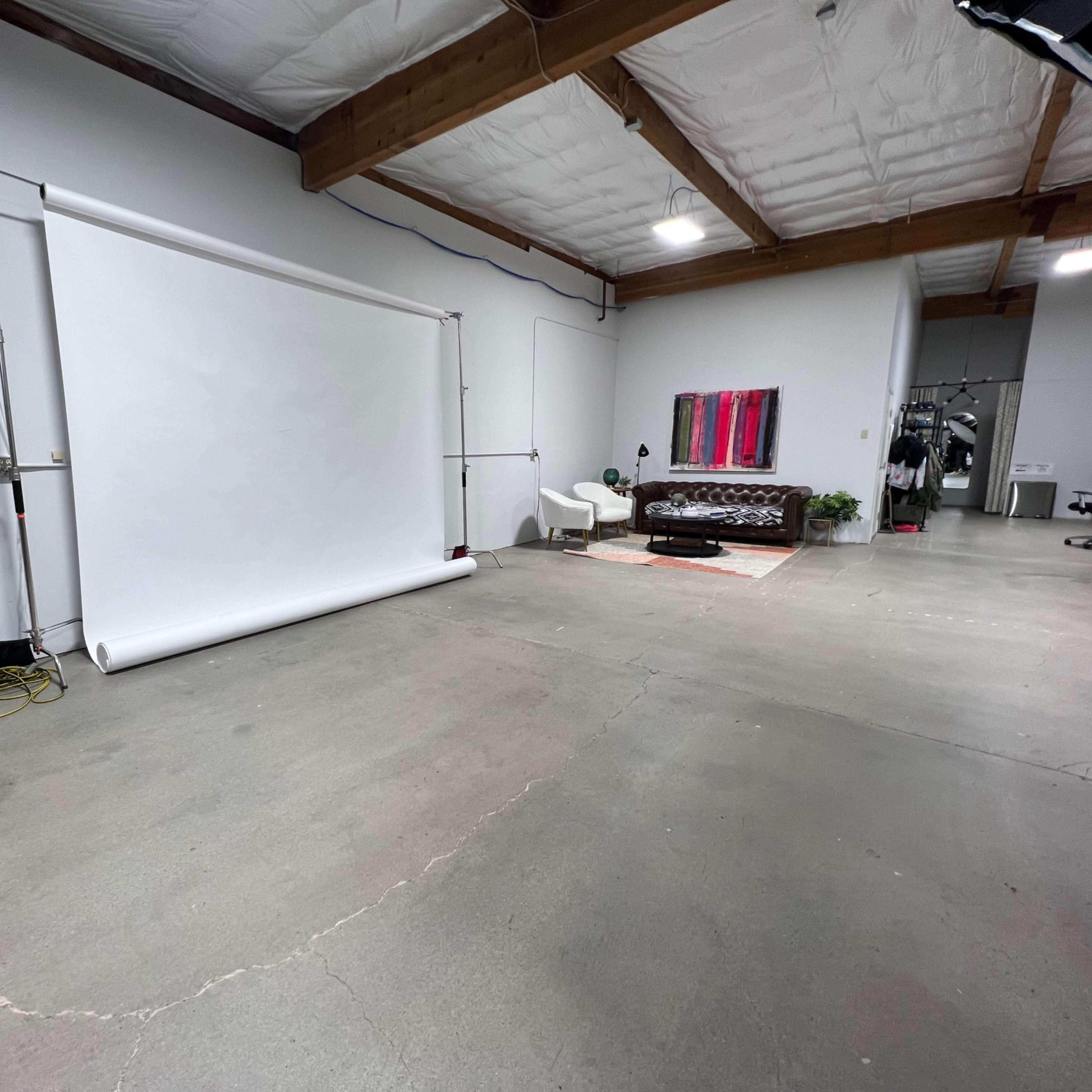 A spacious studio with a roll-up backdrop, a brown leather couch, and minimal furnishings on a concrete floor.