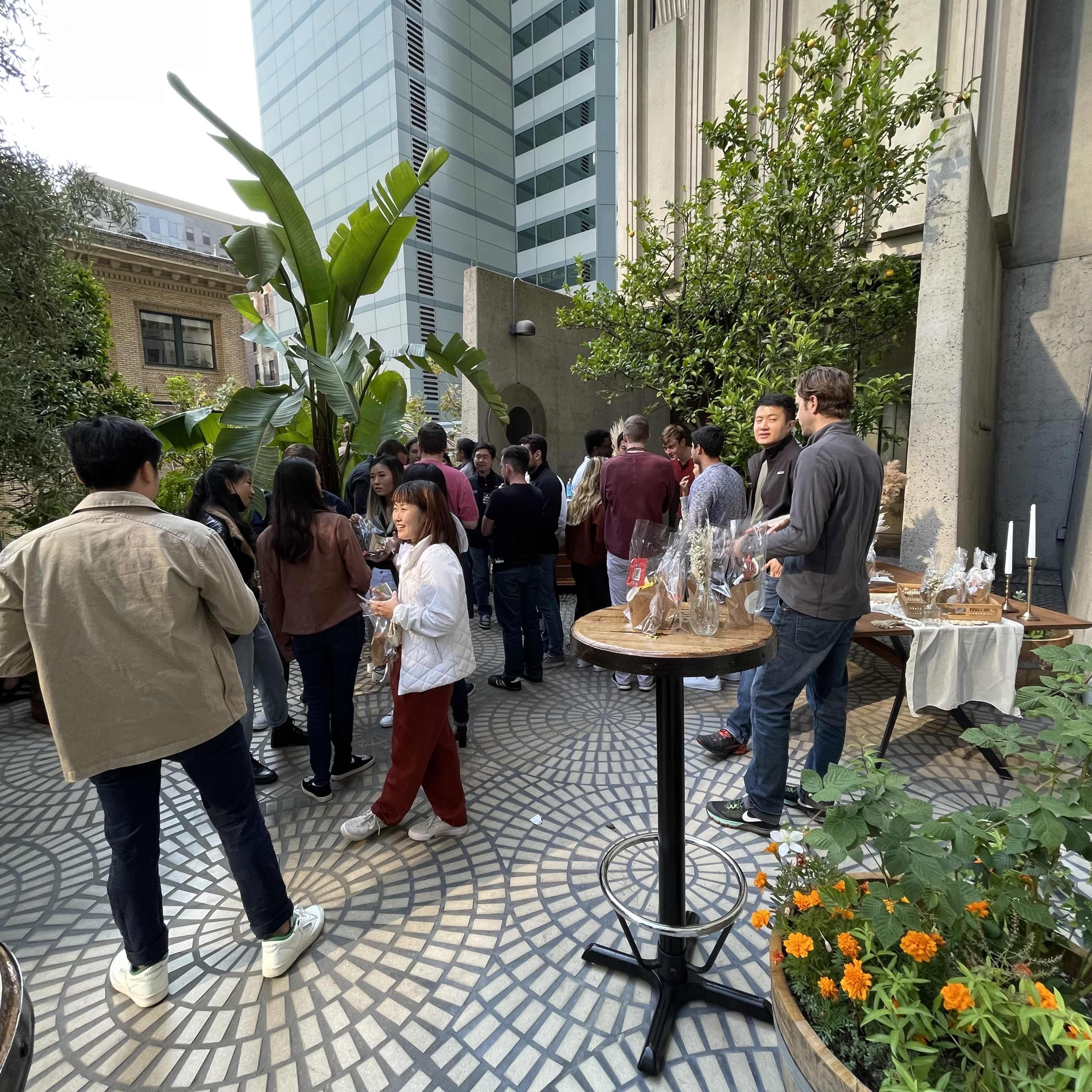 A group of people gathers on a patio surrounded by greenery and plants, with a table displaying items in the background.
